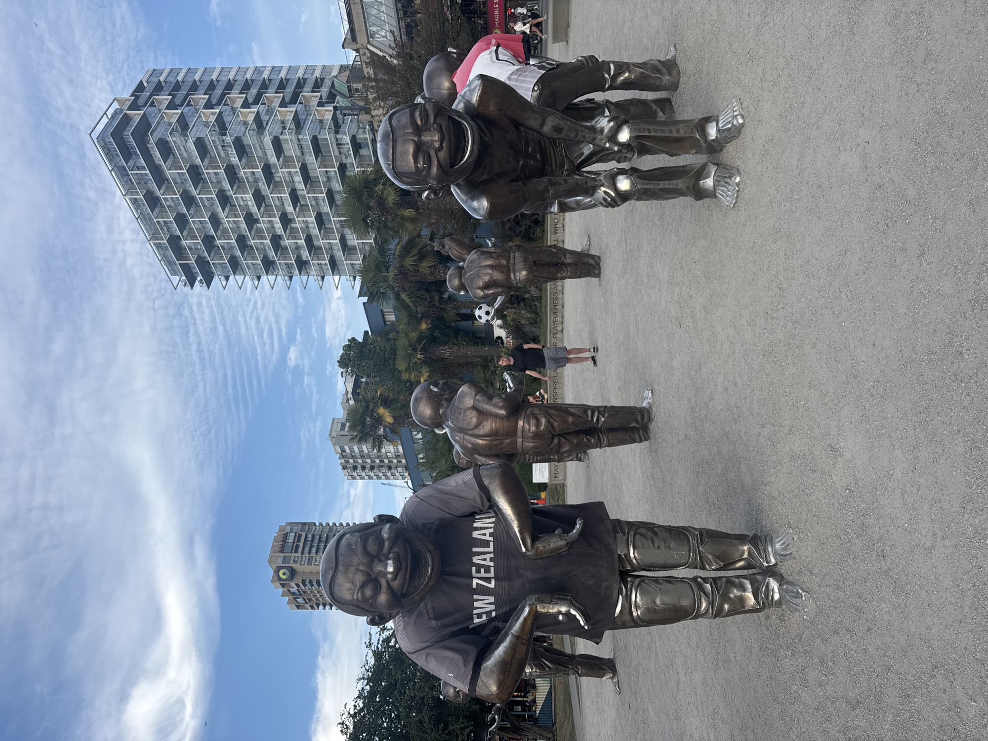 In the foreground of the image, several bronze statues of laughing figures are arranged on a gravel surface. The largest statue in the center wears a dark grey t-shirt with "NEW ZEALAND" printed in white letters. Behind them, other statues are visible, some holding a soccer ball. A man in a black t-shirt and grey shorts stands in the middle ground, looking towards the statues. In the background, several modern high-rise buildings are present, along with palm trees and a partially visible white truck. The sky is blue with scattered white clouds. This scene is located in the public art installation known as "Laughing Stats" in the Jack Poole Plaza, Vancouver, Canada.