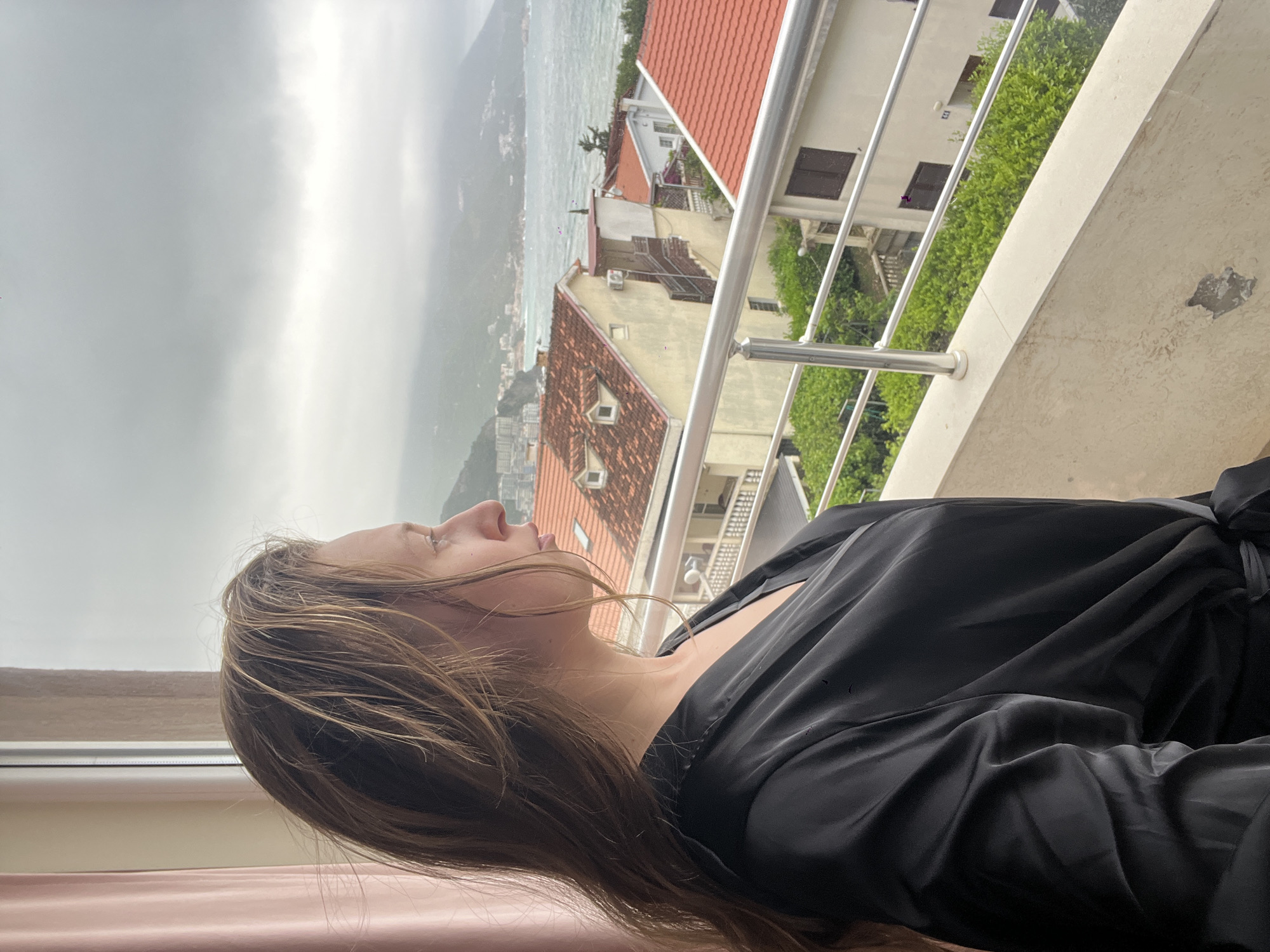 A woman with long brown hair is standing on a balcony, looking up at the cloudy sky. She is wearing a black silk robe. Behind her are buildings with red tile roofs, a metal railing, and some greenery. In the distance, there is a body of water and mountains. The location is likely Bečići, Montenegro, based on the architecture and coastal landscape.
