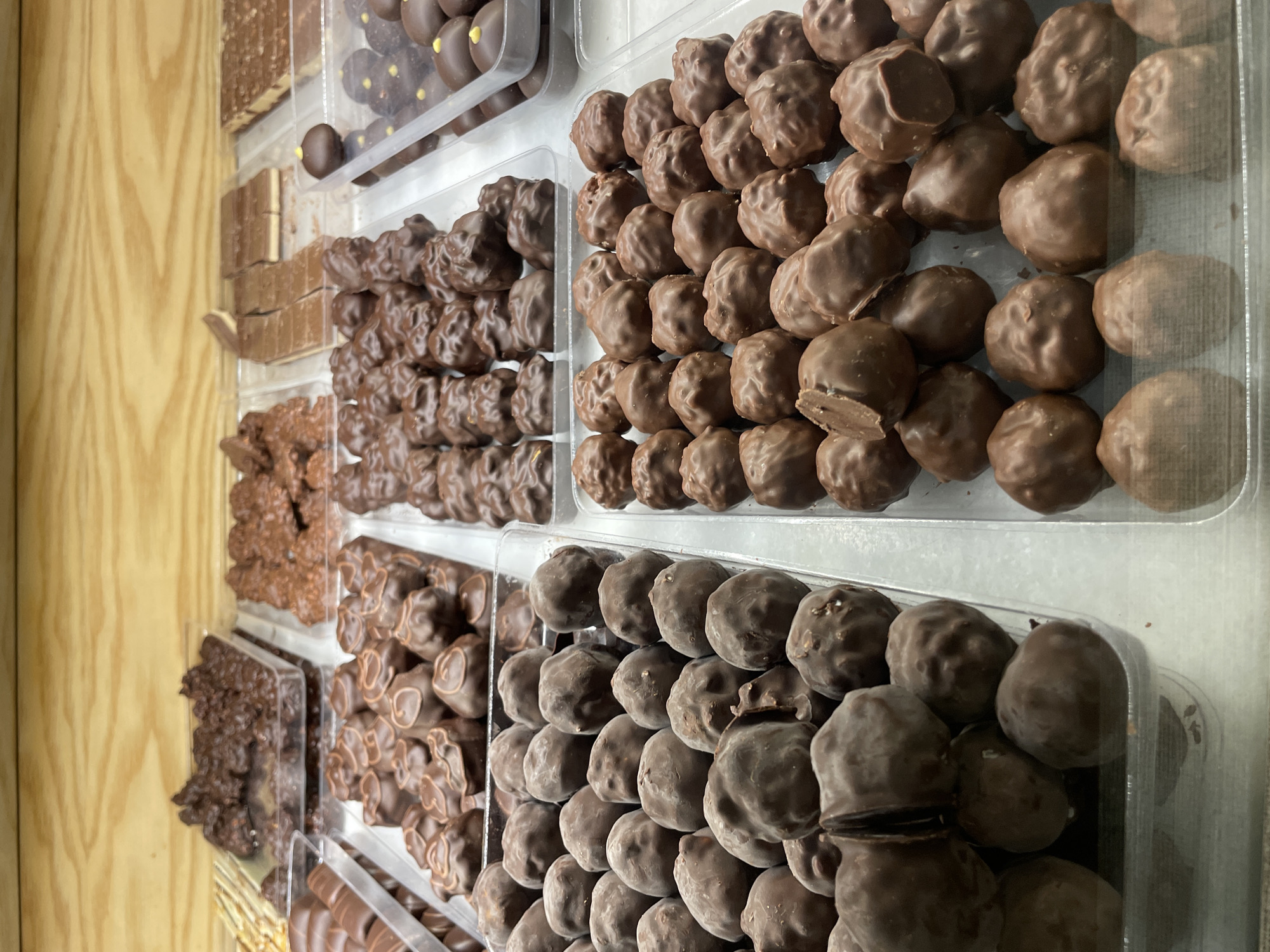 The image displays a selection of chocolate candies arranged in clear plastic containers. The candies vary in shape and type, including chocolate bars, clusters, and round truffles, all presented neatly in rows. The setting appears to be indoors, possibly a confectionery shop or a display at a market, evidenced by the wooden paneling in the background.
While I cannot confirm the location in Buenos Aires, Argentina, the presence of diverse chocolate candies in an organized display suggests a retail environment.