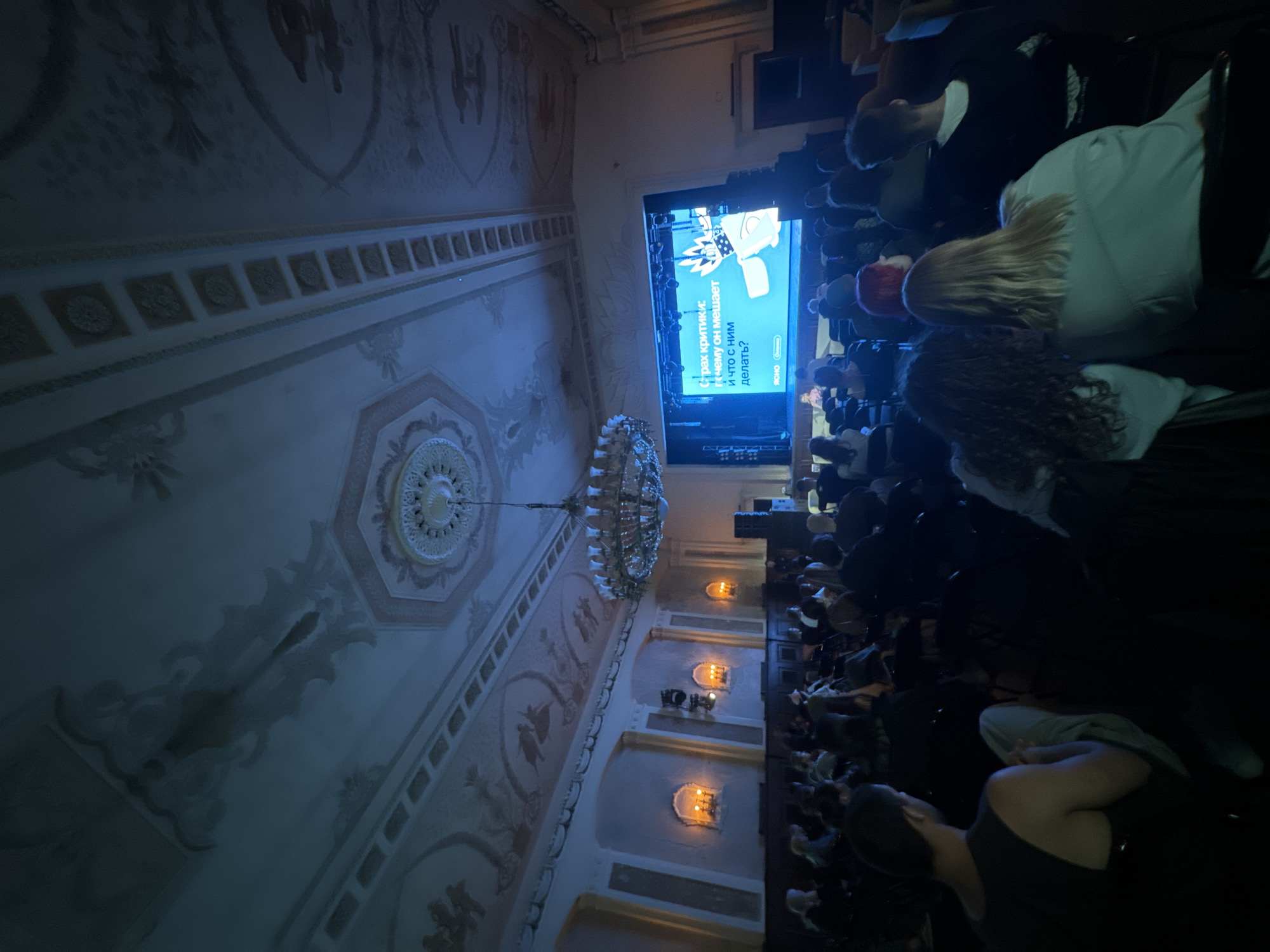 A wide-angle, eye-level, interior shot depicts an audience seated in a dimly lit, ornate hall, facing a brightly lit stage. The hall features decorative plasterwork on the ceiling and walls, with large arched alcoves containing lamps casting a warm glow. A large, multi-tiered chandelier hangs from the ceiling. The audience, comprised of approximately 50-70 individuals, is seated in rows of dark chairs. Many are facing the stage, with some engaged with their phones. The stage is dominated by a large screen displaying text in Russian, which reads "Страх критики: почему он мешает и что с ним делать?". There are speakers and other stage equipment visible. The overall atmosphere is that of a presentation or lecture being held in a historical venue. The image does not contain enough information to determine the specific location in Moscow, Russia.