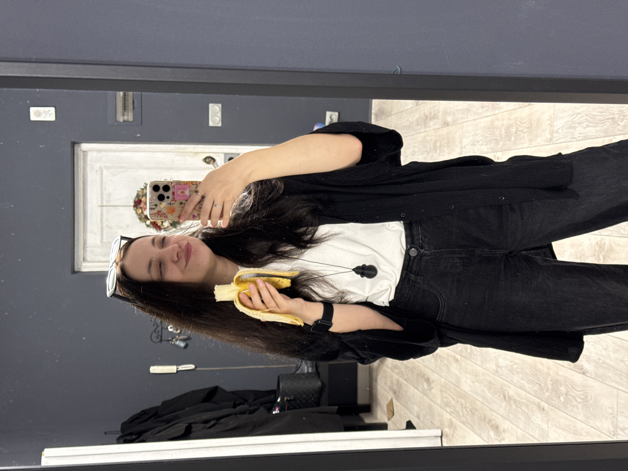 A woman with long dark hair and glasses perched on her head stands in front of a dark grey wall, holding a partially peeled banana in her right hand and a floral-patterned smartphone in her left. She is taking a selfie in a mirror, with her eyes closed and a playful expression on her face. She is wearing a white t-shirt, a black open-fronted shirt, and black jeans. A black smartwatch is visible on her left wrist. In the reflection of the mirror, a white door with a wreath on it and a light-colored wooden floor can be seen. The setting appears to be indoors, likely a home or dressing room.