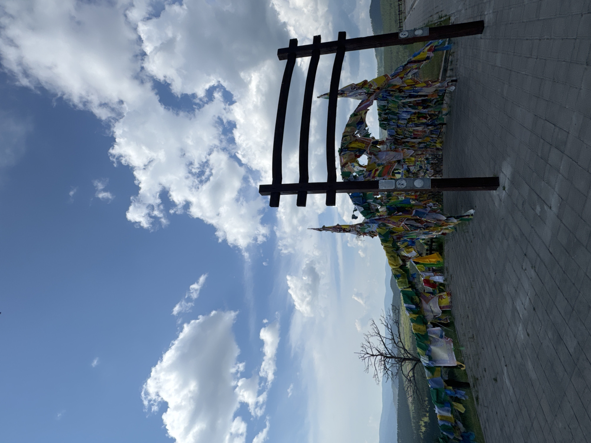 The image shows a pathway in a scenic outdoor location with a clear blue sky dotted with white clouds. In the foreground, a paved pathway made of grey interlocking stones leads towards a large wooden archway adorned with numerous colorful prayer flags. The flags, in shades of blue, green, yellow, red, and white, are strung across the arch and extend along a railing to the left, creating a vibrant canopy.
Behind the archway, a treeless landscape unfolds with rolling hills covered in green vegetation and distant mountains under a hazy sky. To the left of the arch, a bare, skeletal tree stands against the sky, its branches stark and distinct. The overall impression is one of a spiritual or cultural landmark situated in a natural, elevated setting. The location is likely a viewpoint or a place of religious significance. Based on the distinctive prayer flags and the mountainous terrain, this could be a location associated with Buddhism, potentially within the region of Buryatia, Russia, given the mention of Ulan-Ude.