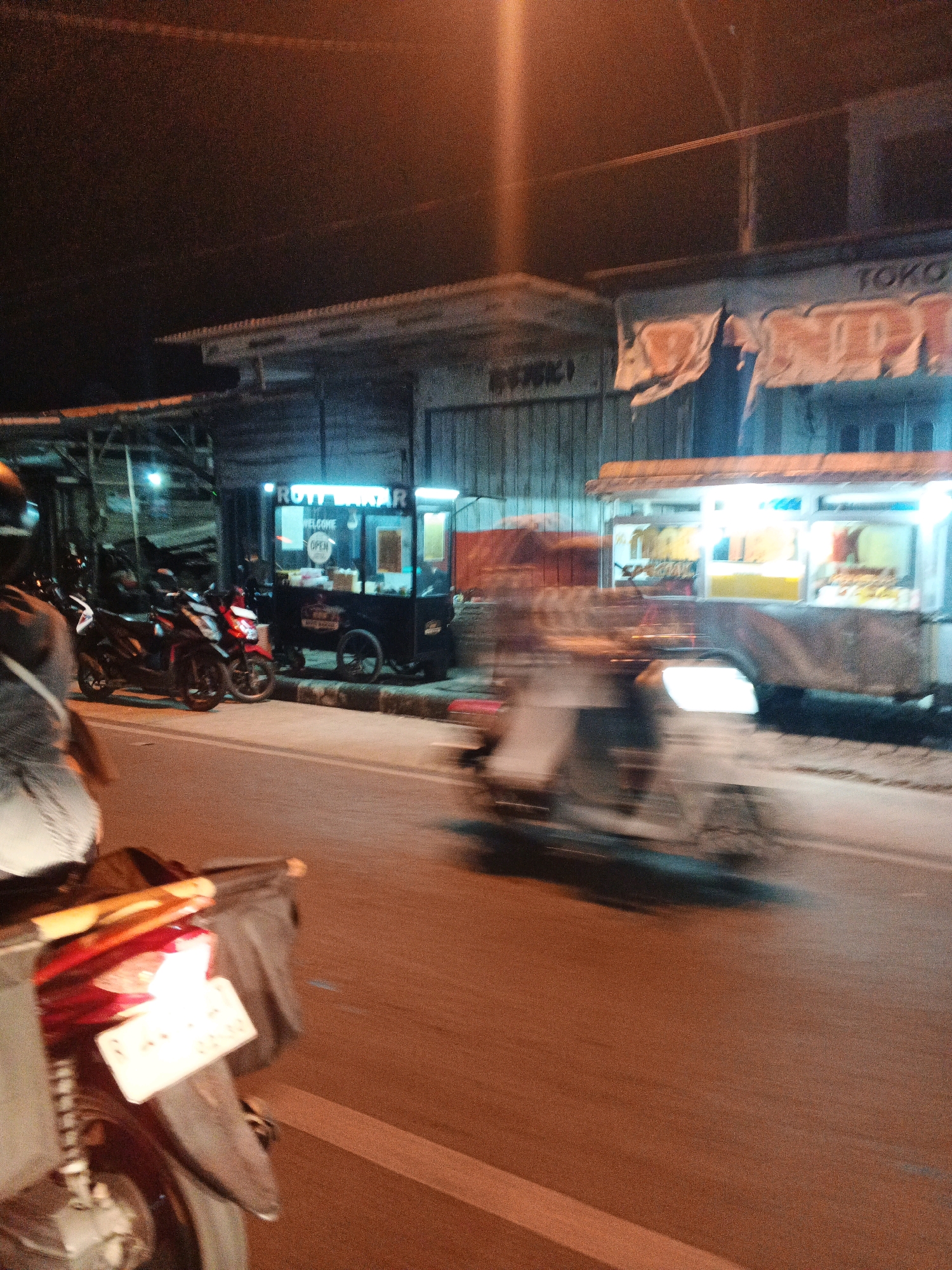 A street scene at night captures a food stall with neon lights, selling "ROTY BAKAR" (grilled bread). Several motorcycles are parked near the stall, and a person on a moving motorcycle is in the foreground, slightly blurred. Another blurred motorcycle is passing by on the street. The background shows a building with a sign that reads "TOKO". The image has a grainy texture and a warm, orange hue from the streetlights.