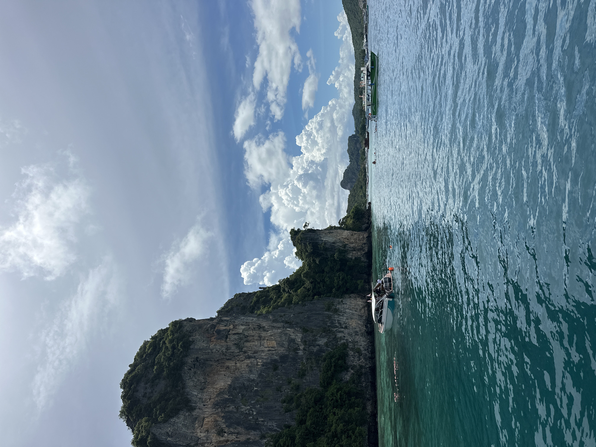 A group of boats are seen floating on the turquoise waters of Ao Nang, Thailand. A large, rocky island covered in lush green vegetation dominates the left side of the frame, with its sheer cliffs rising dramatically from the sea. Several people are visible on the boats and swimming in the water, enjoying the tropical scenery. The sky is partly cloudy, with large white cumulus clouds scattered across a bright blue expanse. In the distance, more islands and boats can be seen, suggesting a popular tourist destination. The water is clear and calm, with gentle ripples reflecting the sunlight.