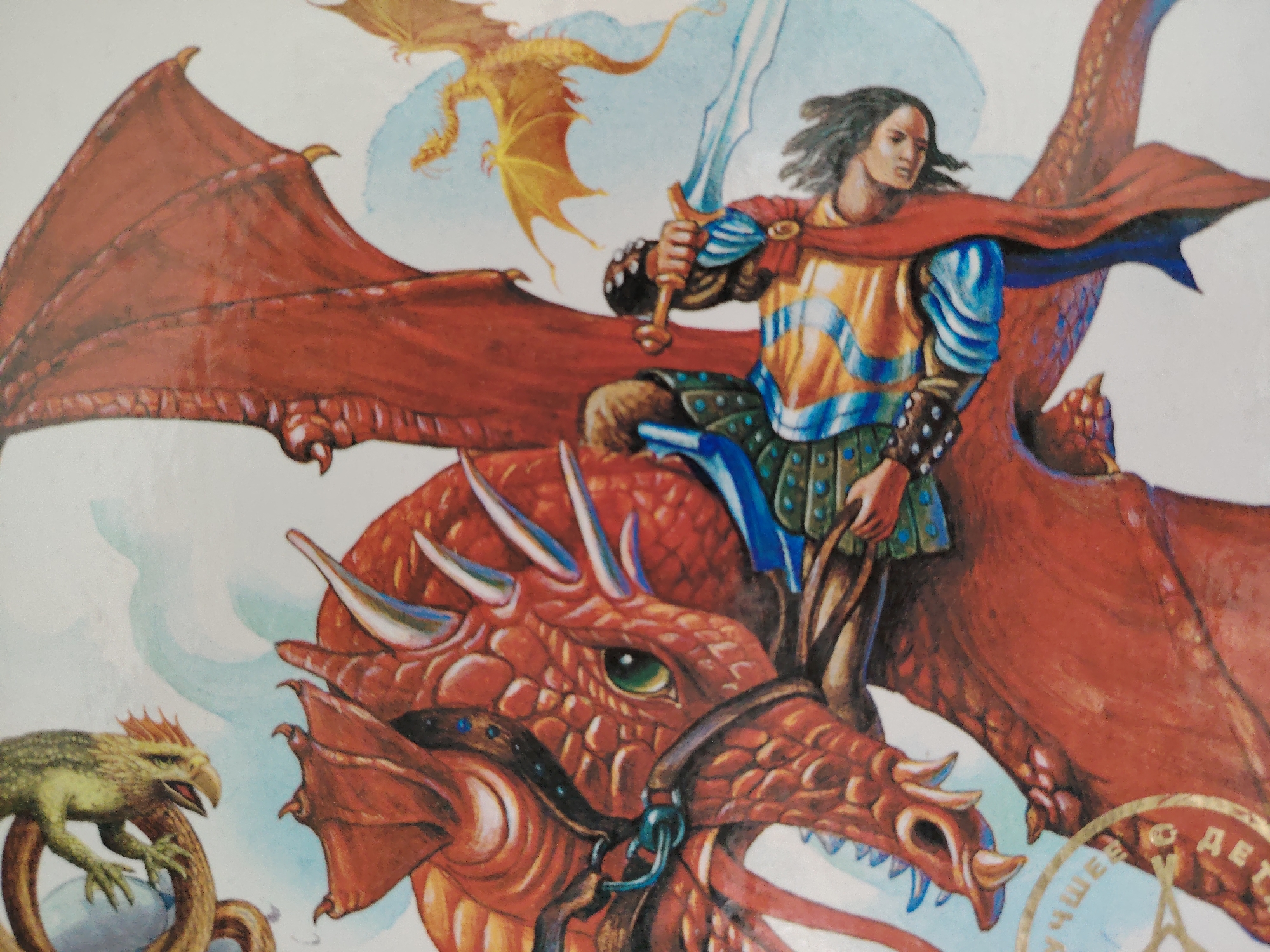This is a watercolor illustration of a fantasy scene. The main subject is a young man with long, dark hair, wearing a red cape, a golden and blue tunic, and green armor. He is holding a silver sword aloft and appears to be riding a large, red dragon. The dragon is depicted with its mouth open, sharp teeth, and green eyes, and it has large, leathery red wings. In the background, a smaller golden dragon flies in a pale blue sky, and a small green creature resembling a lizard or wyrm is visible on the left side of the image, perched on a coiled brown object. The overall impression is one of action and adventure, with the rider and dragon appearing to be in mid-flight or battle. The style is reminiscent of classic fantasy art, with vibrant colors and detailed rendering of the creatures and rider's attire. There is some visible text in a circular golden seal in the lower right corner, appearing to be in Cyrillic script, possibly indicating a publisher or origin. The text is not fully legible but seems to include letters like "ДЕТ" and "ЩЕ". The setting is a fantastical, possibly mythical realm, implied by the presence of dragons and the knightly attire of the rider. There are no clear indicators of time of day or specific weather conditions beyond the pale blue sky. The emotion conveyed is one of heroic action and perhaps a touch of menace from the dragon's open mouth.