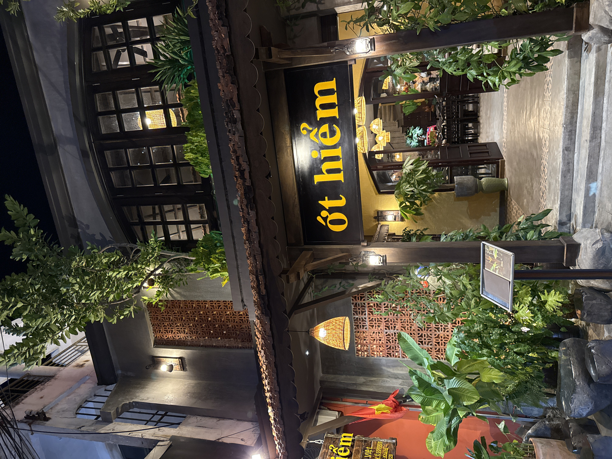 This is a night-time outdoor shot of a restaurant entrance in Nha Trang, Vietnam. The building has a traditional Vietnamese architectural style, with a tiled roof overhang and decorative terracotta screens. Lush green plants are abundant around the entrance, creating a tropical ambiance. A large, dark sign with yellow lettering that reads "ớt hiểm" (meaning "chili" in Vietnamese) is prominently displayed. Inside, several woven pendant lights illuminate tables and chairs. The scene is well-lit by multiple lamps and the interior lights of the restaurant.