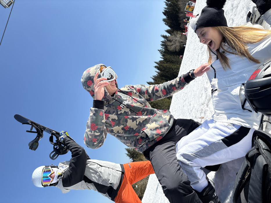 The image depicts three individuals on a snow-covered slope under a clear blue sky, located in a ski resort near Bistritsa, Bulgaria.
In the background, a line of dense pine trees defines the edge of the slope. A partial view of a ski lift chair is visible in the upper right.
Three subjects are present:
1. **Subject 1 (Left):** A person standing upright, facing right, wearing a white ski helmet with iridescent goggles, a grey and black ski jacket, and bright orange ski pants. This individual holds a black snowboard vertically with their right hand, raised above their head.
2. **Subject 2 (Center):** A person in a crouching position, leaning towards Subject 3. This individual wears a hooded camouflage sweatshirt with red rose patterns, black ski pants, and white-framed goggles pushed up onto their forehead. Their left arm is extended downwards towards Subject 3, and their right hand is near their face, holding or adjusting the goggles. A black smartwatch is visible on their left wrist.
3. **Subject 3 (Right):** A person lying supine on the snow, facing upwards and to the left, with an open mouth suggesting laughter. This individual wears a white ski suit with black and grey accents and a black knit beanie with a visible pom-pom on their head. Light brown hair is visible beneath the beanie.
Additional objects include a black backpack in the lower left foreground and a black helmet with red accents near Subject 3 in the lower right foreground. The interaction appears playful.