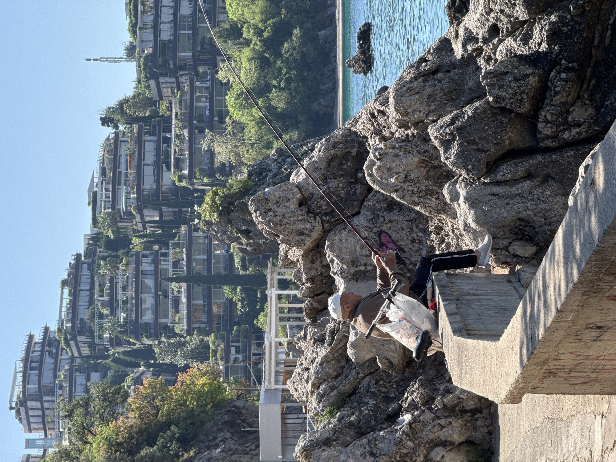 An eye-level outdoor shot depicts a man fishing from a concrete structure adjacent to rocky terrain. The man, wearing a hat, jacket, and dark pants, sits with his legs extended, one foot bearing a white shoe. He holds a fishing rod, and a plastic bag rests near him. The background includes a cityscape with multiple multi-story buildings and trees, and a body of water is visible as well. The scene is located in Budva, Montenegro.