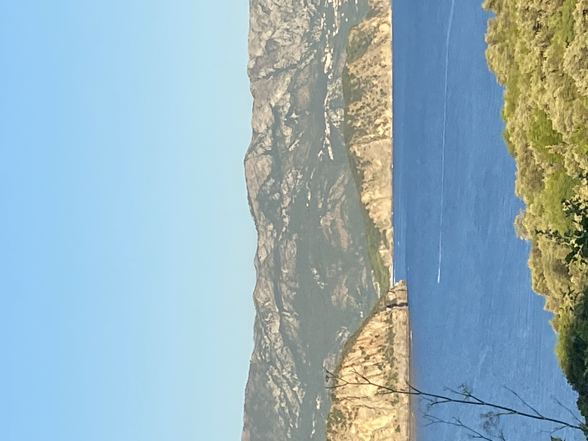 A landscape photograph captures a view of the Adriatic Sea with rugged, mountainous terrain and a clear blue sky. In the foreground, a patch of dense green foliage with some bare branches is visible. The middle ground showcases the deep blue sea, with a small white boat leaving a faint wake. Two rocky islands, characterized by their golden-brown cliffs and green vegetation, jut out into the sea. The background is dominated by a range of imposing mountains, their slopes a mix of dark green and light grey rock, with scattered white structures, possibly buildings, visible on the lower slopes. The sky is a pale, cloudless blue, transitioning to a lighter hue towards the horizon. The scene suggests a coastal view in a Mediterranean or Balkan region, likely within Dubraljevina, Montenegro, given the characteristic coastline and mountainous backdrop.