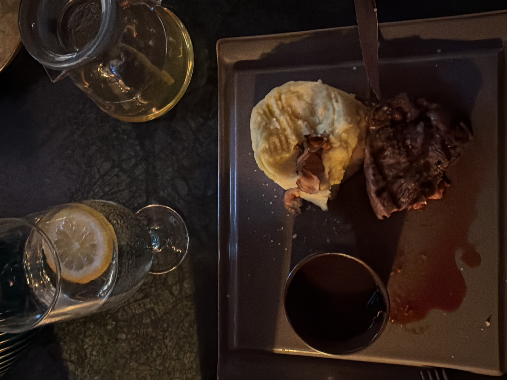 A high-angle shot displays a meal on a square plate, situated on a dark, patterned surface. The plate contains a serving of mashed potatoes topped with what appears to be crumbled bacon, a piece of grilled meat with visible grill marks, and a small bowl of dark sauce. A splatter of reddish liquid is also present on the plate. To the left, a clear glass pitcher filled with a yellowish liquid rests near a wine glass containing a lemon slice. Utensils are partially visible at the bottom and left edges of the frame. The location appears to be a dimly lit restaurant. The city and region are unknown.
