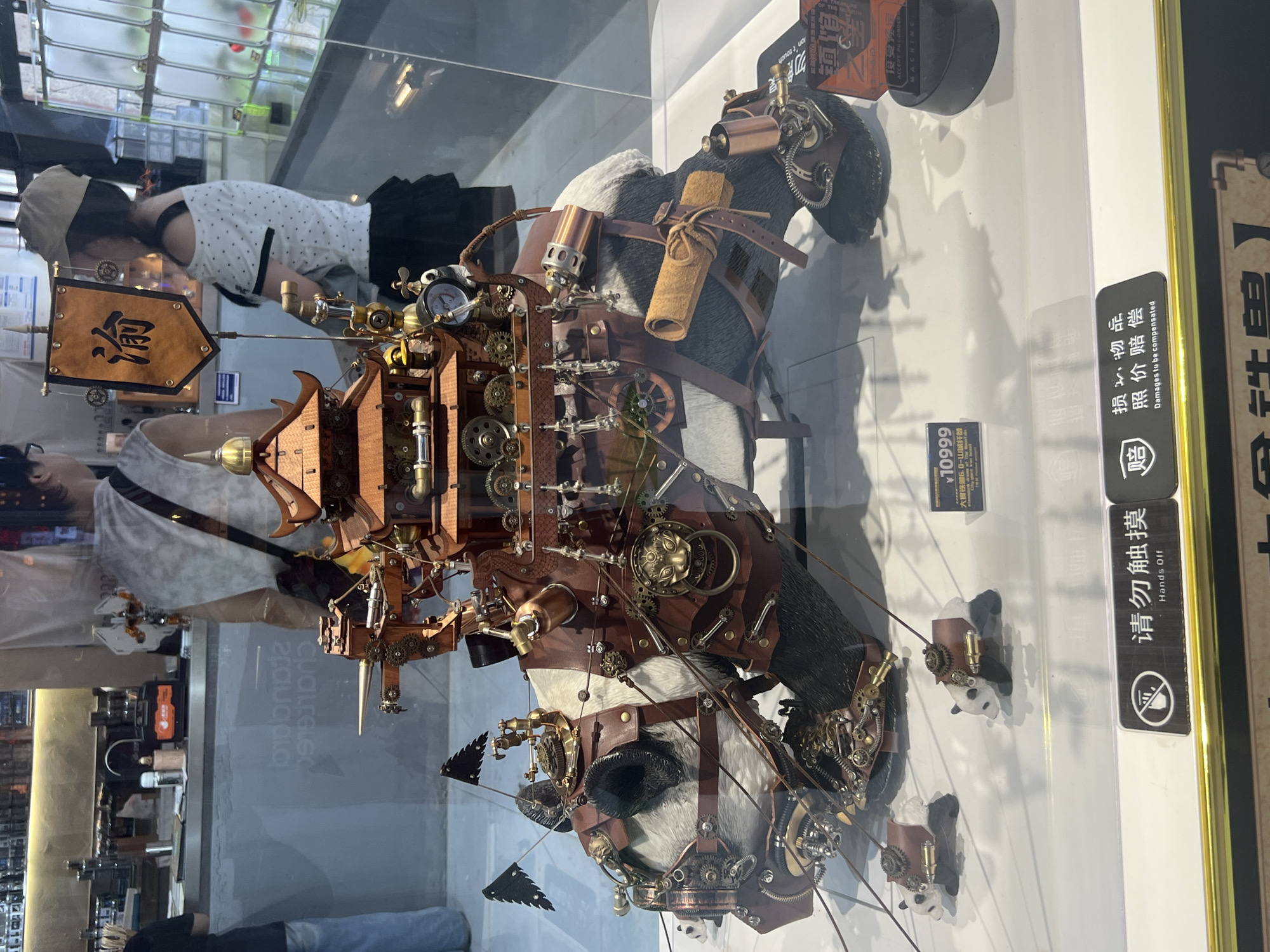 In Yuzhong District, China, a display case exhibits a steampunk-themed diorama featuring a large panda figurine adorned with intricate leather and metallic gears, resembling a war machine. A miniature pagoda-like structure is mounted on its back. Two smaller panda figurines in a similar steampunk style are positioned in front of the main figure. The background reveals a store interior with shelves stocked with various items and people browsing. Signage in Chinese and English indicates "Hands Off" and "Damages to be compensated," suggesting the display is a product for sale.