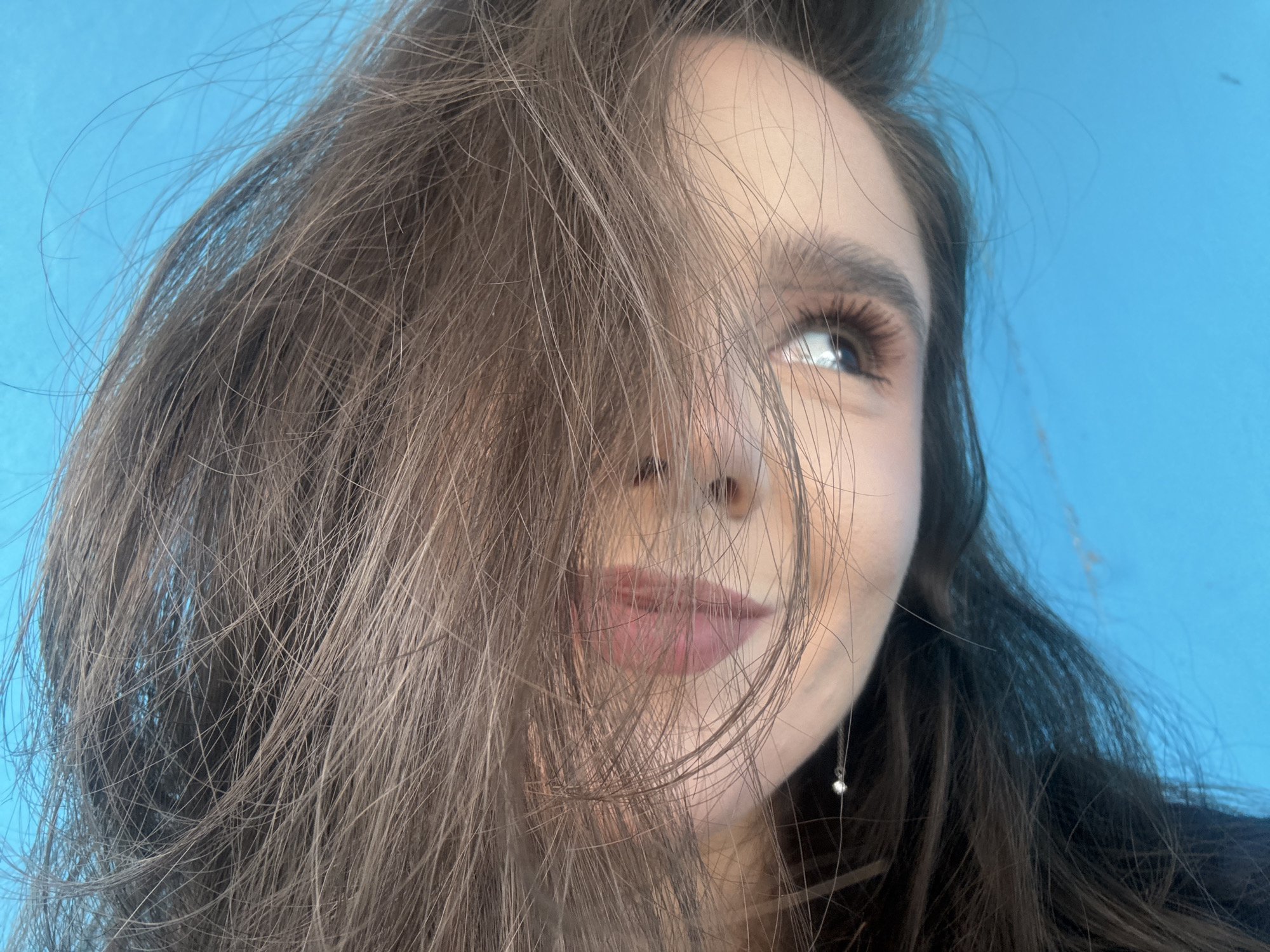 The image is a close-up, medium shot of a young woman's face, taken from a low angle. She has long, brown hair that partially obscures her face, with strands falling over her left eye and across her nose and mouth. Her right eye is visible, looking upwards and to the right, with dark eyelashes and a hint of mascara. Her lips are painted with a soft pink lipstick, and she has a subtle smile. A delicate silver earring with a small pearl or gem is visible hanging from her right ear. The background is a vibrant, solid blue. The lighting suggests it is daytime, possibly outdoors due to the natural light reflecting in her eye, or in a well-lit indoor space. The overall mood conveyed is one of casualness and slight playfulness. There is no visible text in the image. The location context provided is Shulhynka, Ukraine, which does not offer any specific visual cues within the image itself.