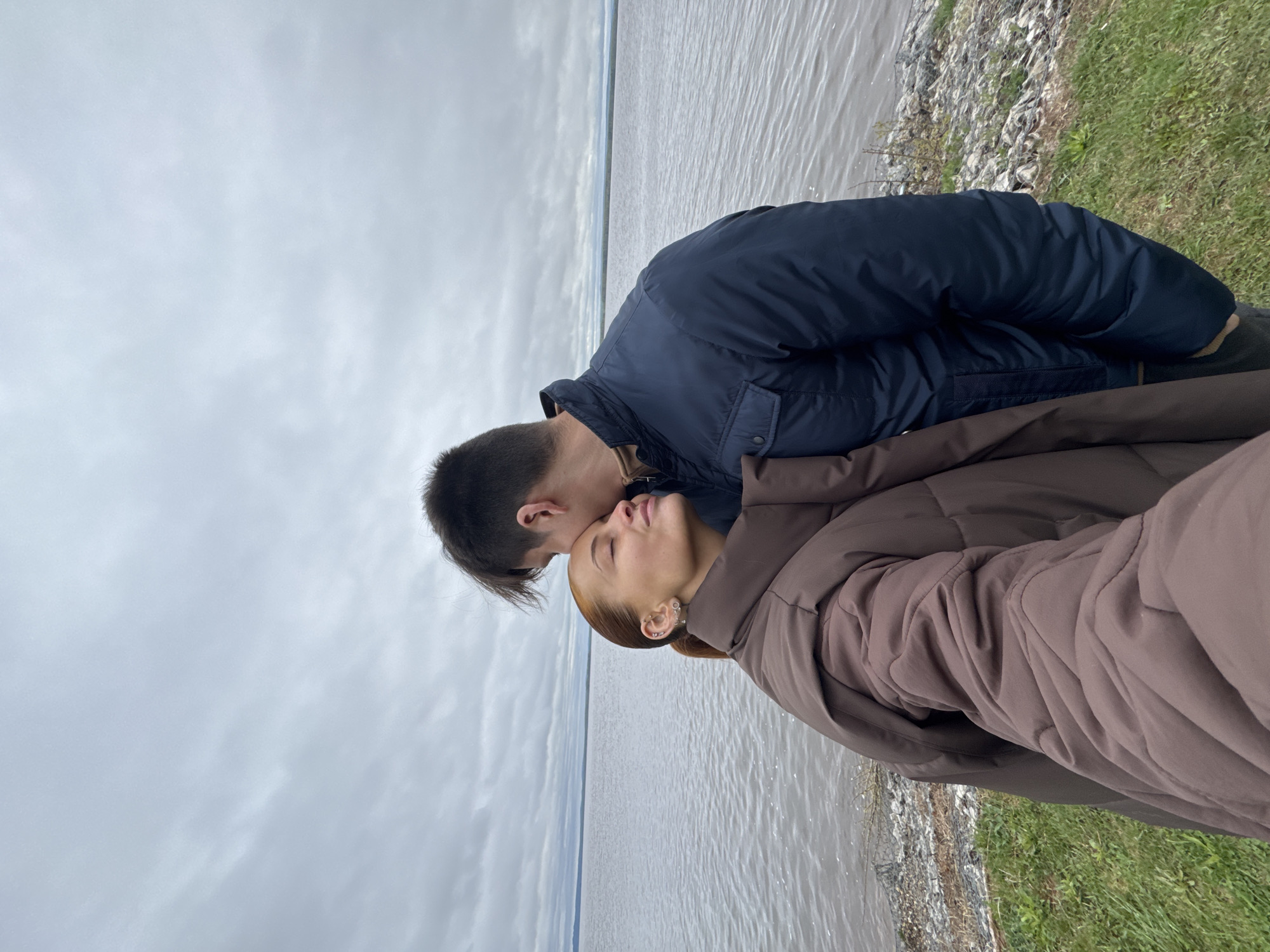 A photograph shows a man kissing a woman on the forehead. The man has short, dark hair and wears a dark blue jacket. The woman has reddish-blonde hair pulled back, earrings, and a brown puffer jacket. They are standing by a body of water, possibly a wide river or lake, with a gray sky overhead. The shoreline has some grass and rocky areas. The location is Chastyye, Russia.
