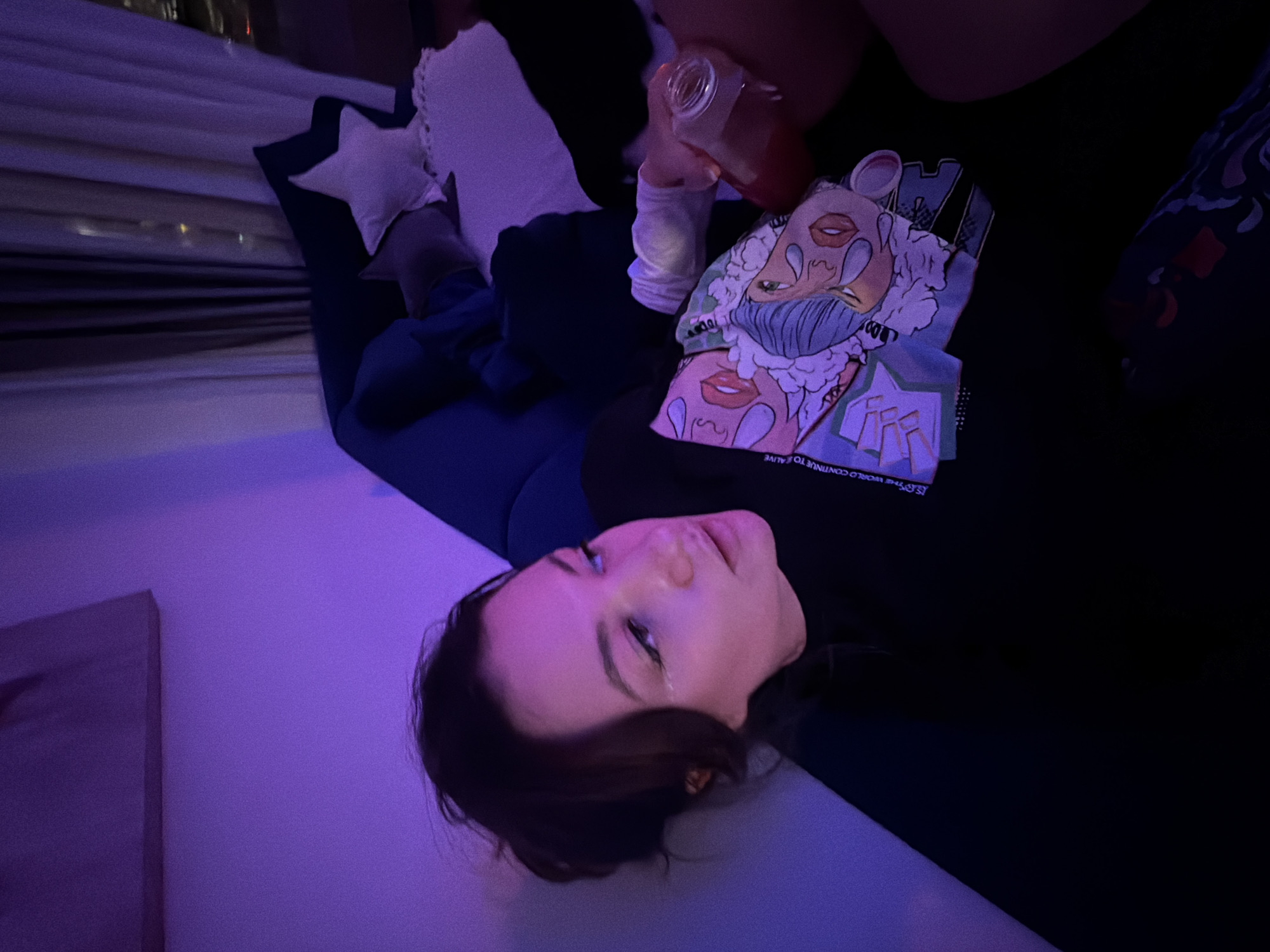 A woman lies on a blue sofa in a room illuminated by purple and blue light. She is wearing a black long-sleeved shirt with a print. The woman holds a glass bottle in her hand. Behind the sofa are decorative pillows, including one shaped like a star. A window shows blurred lights, potentially indicating a city view. The specific location in Belgrade, Serbia cannot be determined from the available information.