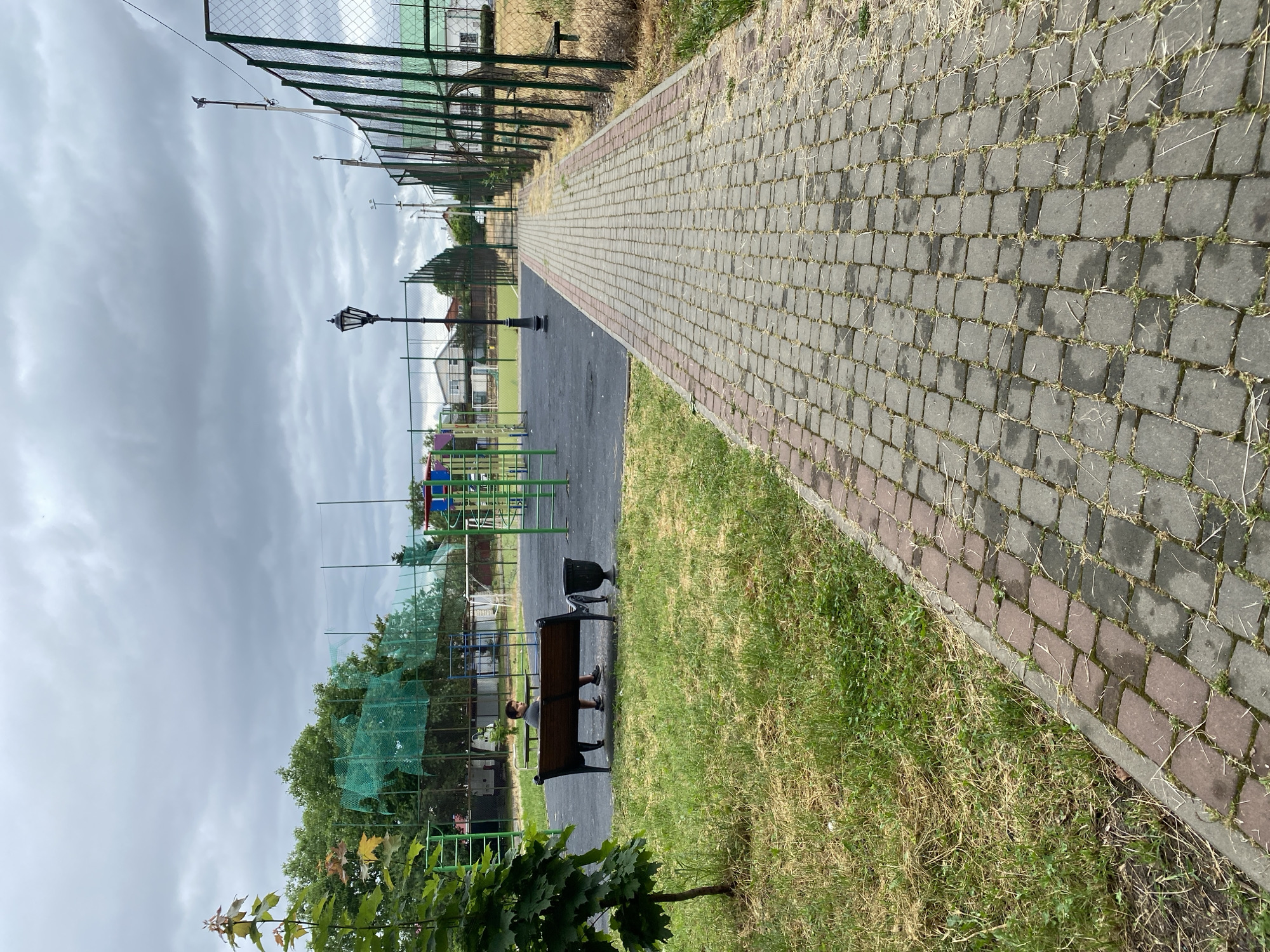 The image depicts a paved walkway in an outdoor public space in Zapadny, Russia. The walkway, constructed of interlocking grey and reddish-brown cobblestones, leads away from the viewer into the distance, bordered on the right by a chain-link fence with green covering. To the left of the walkway, a grassy area is visible, with a young tree in the foreground and a wooden bench with a metal trash receptacle. A person, appearing to be a young male wearing a grey shirt and dark shorts, is seated on the bench. Beyond the bench, a playground area is visible, featuring colorful structures and green netting. Further in the background, a sports field with a fence and some buildings can be seen under a cloudy sky. A single lamppost stands near the playground.
