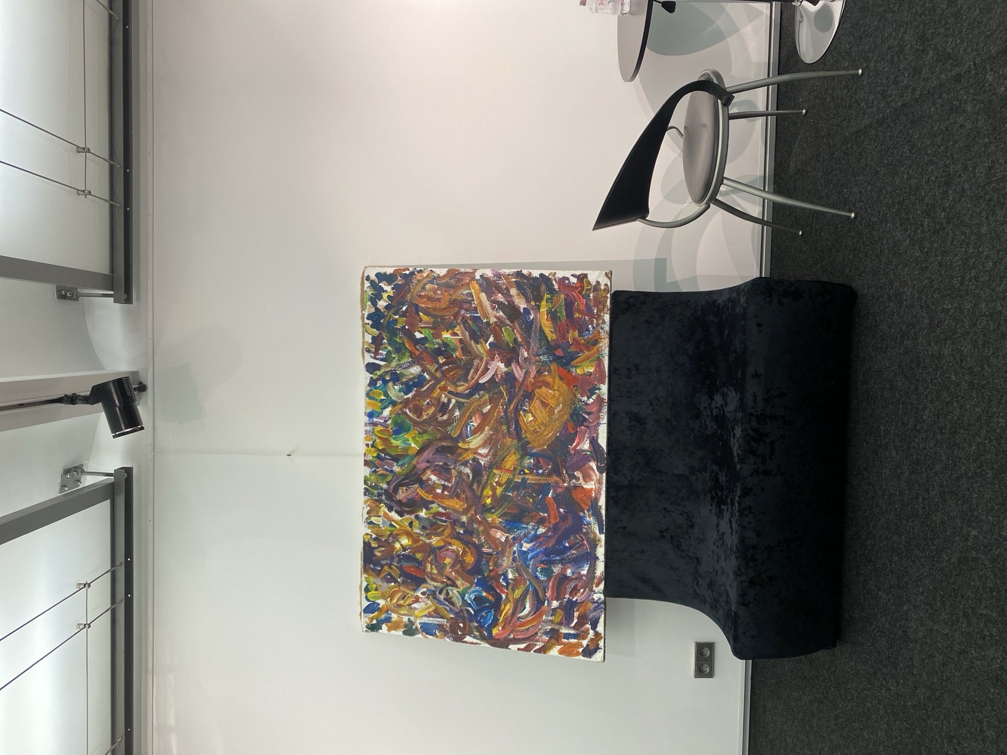 A rotated, eye-level, indoor shot reveals an art installation or exhibit. A colorful, abstract painting hangs on a white wall. A small, dark blue velvet chaise lounge sits beside the painting. Nearby are two modern chairs with metal frames and black or gray seats. To the left of the painting, a track lighting system is mounted on the wall. The environment appears to be a gallery or exhibition space. The presence of the art, seating, and lighting suggests a setting for viewing and appreciating art. Based on the prompt, it is assumed to be in Moscow, Russia.
