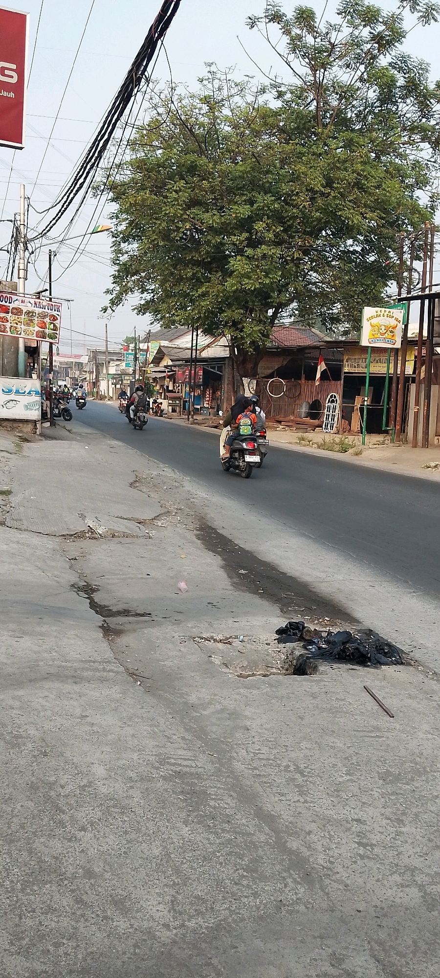 The image depicts a street scene in Satriamekar, Indonesia. A paved road runs through the center, with a concrete sidewalk or shoulder on the left and businesses lining the right. Several motorcycles are visible on the road; one with a rider and passenger is in the foreground, heading away from the camera. Another motorcycle is seen in the middle distance. On the left side of the road, a sign advertising "FOOD & NASI PADANG" is mounted on a pole. Further down the road on the right, there's a sign for "KEDAI SEDULAK & SOP IGA." A large tree with dense foliage is prominently positioned behind the businesses on the right. Electrical wires crisscross the sky. The environment appears to be a commercial or residential street with signs of wear and tear on the pavement, including cracks and a small pile of black trash bags on the ground near the foreground. The sky is clear and appears to be late afternoon or early morning, given the lighting.