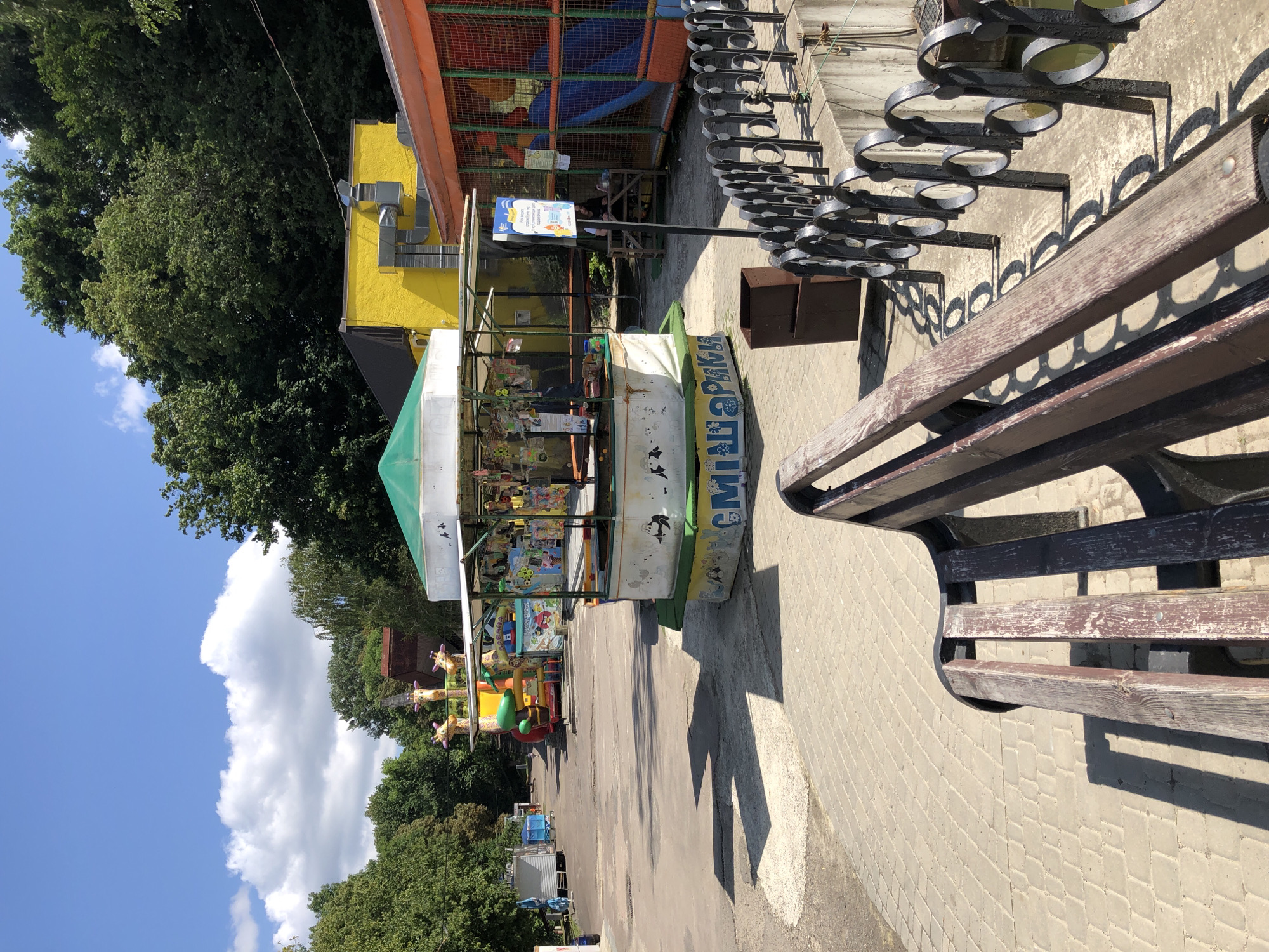 The image depicts an outdoor amusement area on a sunny day. In the foreground, a wooden park bench faces an asphalt path paved with light-colored interlocking stones. To the right, a black metal railing with circular openings runs along a concrete ledge. In the middle ground, a small carousel with a white and green canopy sits empty, adorned with cartoonish figures and colorful decorations. Behind the carousel, a bright yellow building with gray ventilation pipes is visible, and next to it, an orange structure with a netted enclosure and a blue slide suggests a children's play area. In the background, several inflatable attractions, including a giraffe-themed one, are set up, surrounded by lush green trees. The sky is blue with scattered white clouds. The scene is located in the Lviv Vodnograd, an amusement park in Lviv, Ukraine, known for its water rides and family attractions.