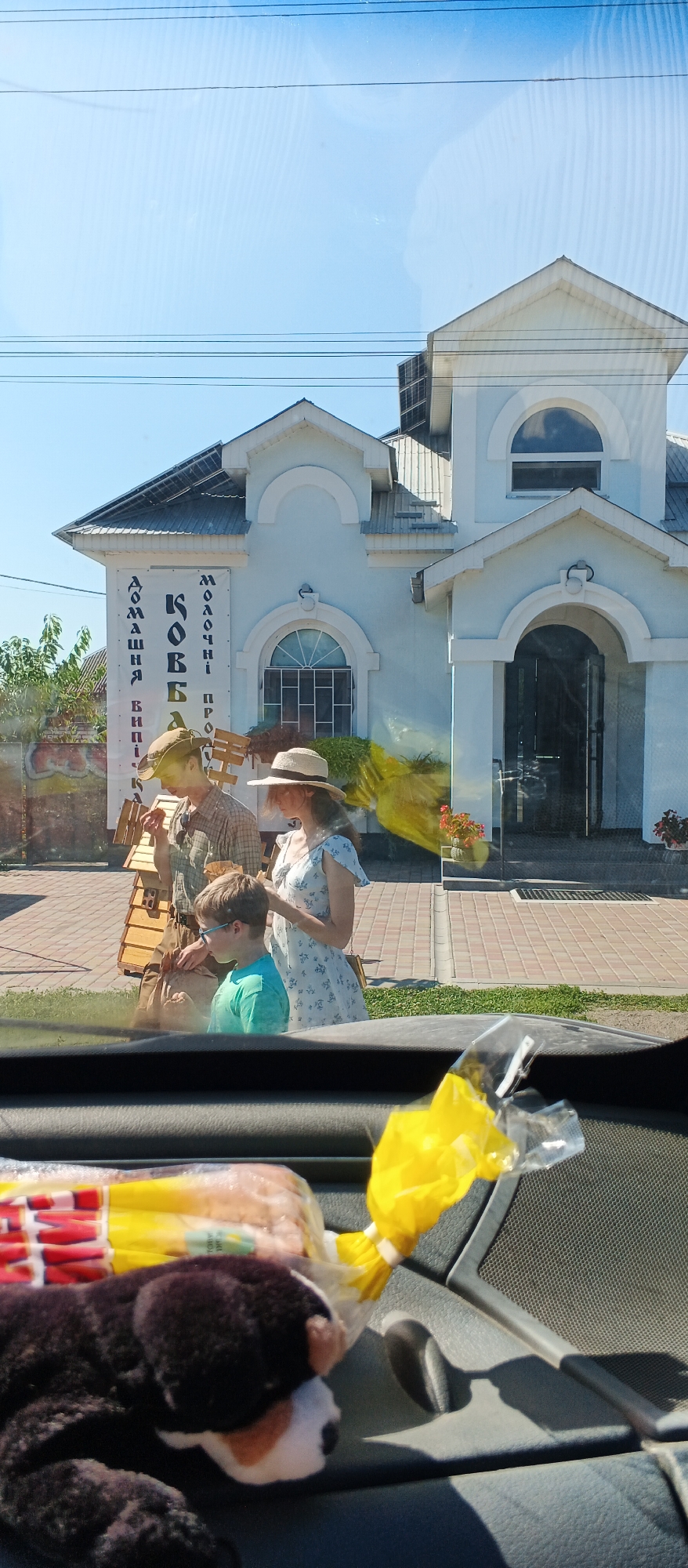 In the city of Ploske, Ukraine, an outdoor scene unfolds in front of a light blue building with white architectural accents. The building appears to be a commercial establishment, with signage visible. A family, comprising a man, a woman, and a young boy, are present in front of the building. The man, wearing a hat and a plaid shirt, is holding a piece of food to his mouth. The woman, also wearing a hat, is dressed in a floral-patterned dress. The boy, in a teal t-shirt and blue-tinted glasses, is looking downwards. In the foreground, a toy dog and a wrapped item, possibly food, are visible on a car's dashboard or interior. The sunlight suggests it is daytime.