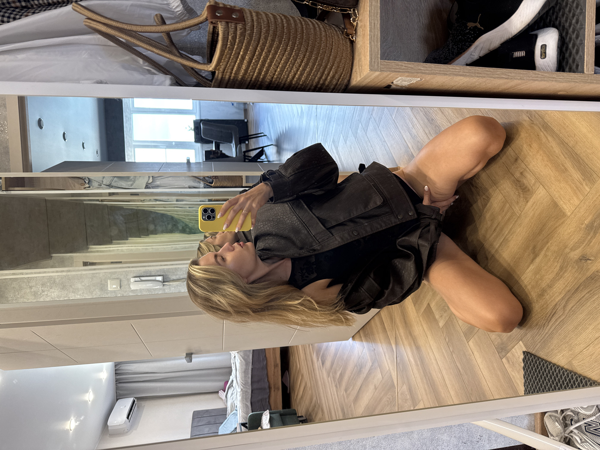 A young woman with blonde hair is kneeling on a wooden floor in front of a full-length mirror, taking a selfie with her yellow smartphone. She is wearing a black tank top and a dark brown faux leather jacket that is unbuttoned and draped off her shoulders. Her knees are on the floor, and her legs are bent, with her right leg positioned in front of her. The mirror reflects the room behind her, which appears to be a bedroom with a bed, an air conditioning unit, and a door leading to another area. To the right of the mirror, a wicker tote bag hangs on a wall, and a wooden shelf unit displays a pair of black and white athletic shoes. The floor is made of light-colored hardwood in a herringbone pattern.