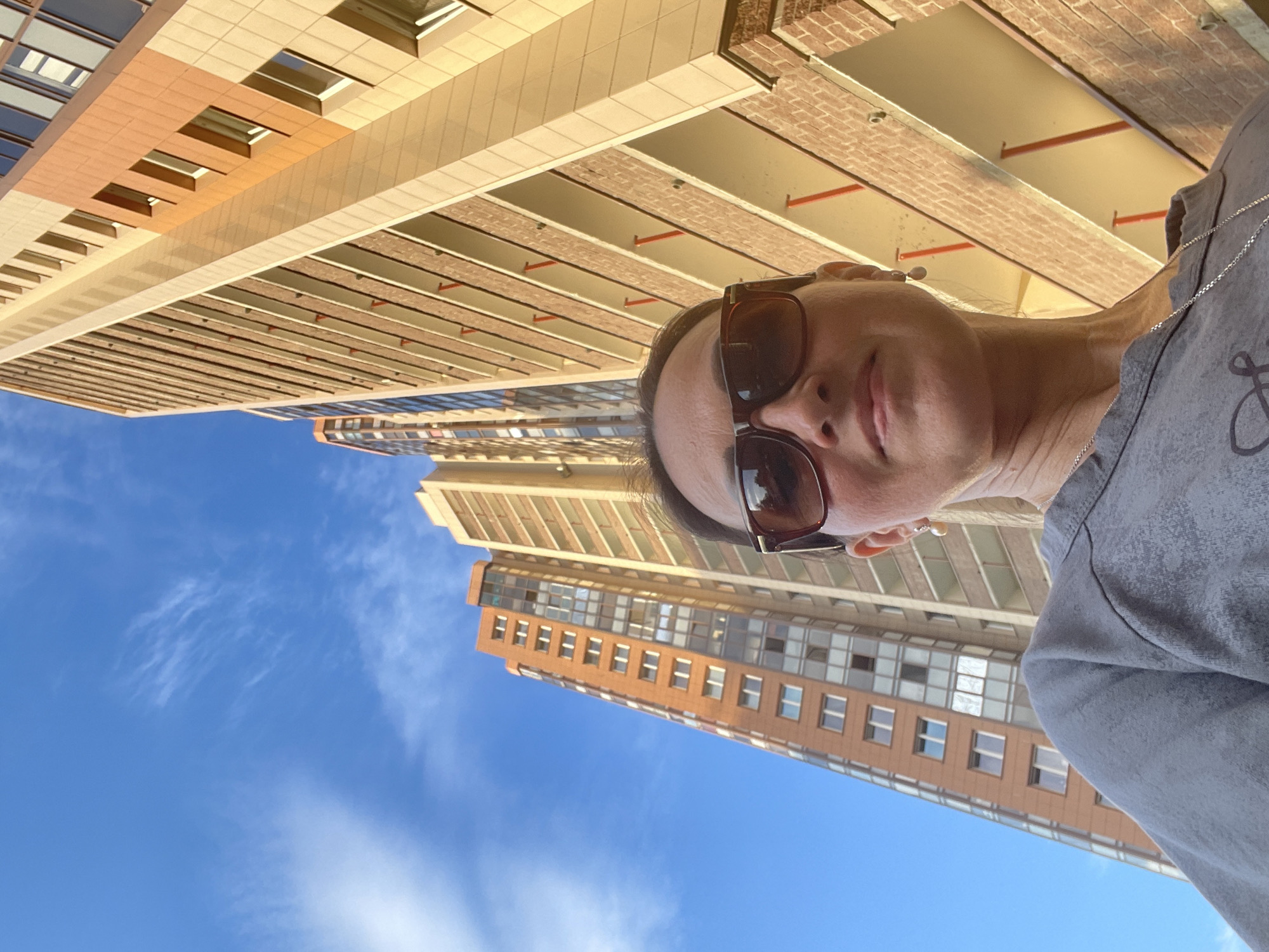In a low-angle, selfie-style shot, a woman with blonde hair and brown-tinted sunglasses stands in front of a bright blue sky with thin white clouds. She is wearing a grey sweatshirt and a silver chain necklace, with a pearl earring visible in her right ear. Her head is slightly tilted upwards. Behind her, the towering facade of a modern apartment building, characterized by rows of balconies and a mix of orange and beige cladding, dominates the background. Another similar, taller building is visible to the left, also under the blue sky. The lighting suggests it is either early morning or late afternoon, casting a warm glow on the building's exterior. The image appears to have been taken outdoors in an urban environment in Saint Petersburg, Russia, possibly in a newly developed residential area.