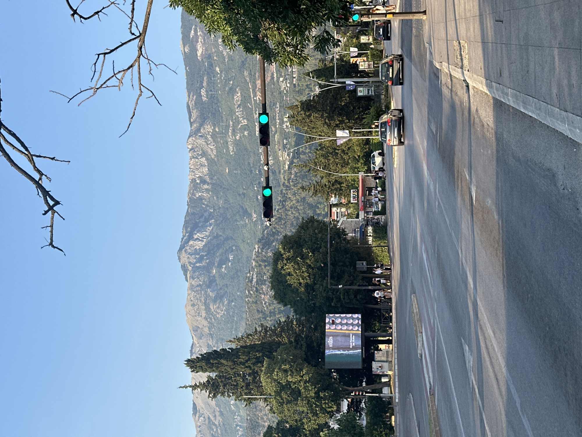 The image captures a street scene in Budva, Montenegro, under a clear blue sky. In the foreground, a multi-lane road with white lane markings is visible, with cars driving on it. A pedestrian crossing is present on the left side of the road. Sidewalks run along the right side of the road, with a few people standing and walking. A large digital billboard displaying advertisements is situated on the left side of the road, surrounded by lush green trees and tall cypress trees. Traffic lights, with green signals illuminated, are suspended over the intersection. In the background, a panoramic view of a mountain range, dotted with buildings, dominates the landscape. The overall scene depicts a bustling urban environment with a backdrop of natural beauty.