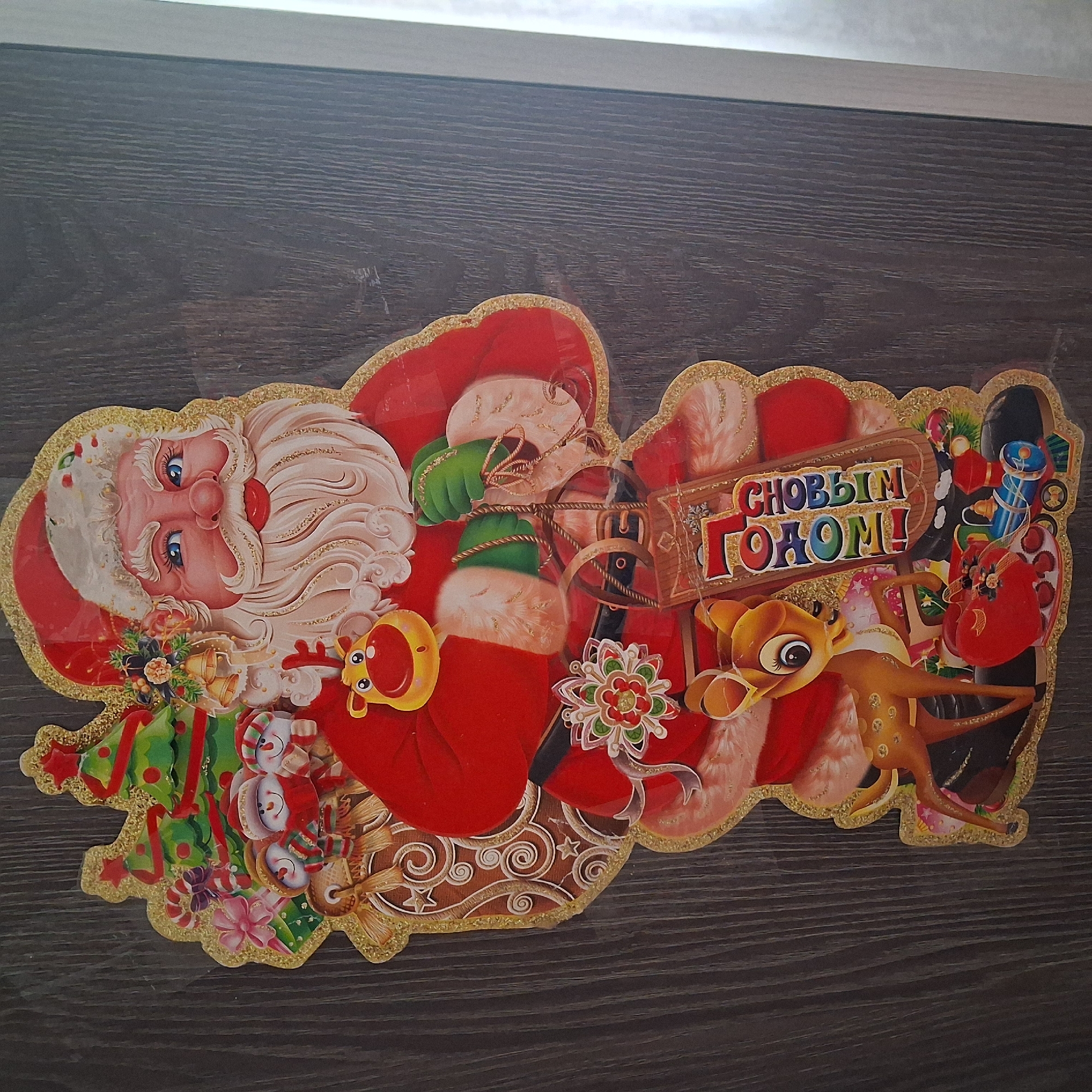 The image displays a flat, festive Christmas decoration of Santa Claus with a sack of gifts. He is depicted with a large white beard and mustache, blue eyes, and rosy cheeks, wearing his iconic red suit trimmed with white fur. To his left, a miniature Christmas tree adorned with ornaments and a red star is visible, along with three snowmen and a wrapped gift. Santa holds green-gloved hands, and his sack appears to be made of burlap with gold glitter accents.
A small reindeer with a yellow face and brown spots stands near Santa's feet, accompanied by a red heart-shaped gift, a toy train, and other festive items. A wooden tag hanging from Santa's sack reads "С НОВЫМ ГОДОМ!", which translates to "Happy New Year!" in Russian. The decoration is affixed to a dark wooden surface, likely a door or wall, characterized by vertical grain patterns. The lighting suggests an indoor environment. The image does not provide any specific details about the city of Bugulma, Russia, or any activities taking place within it.