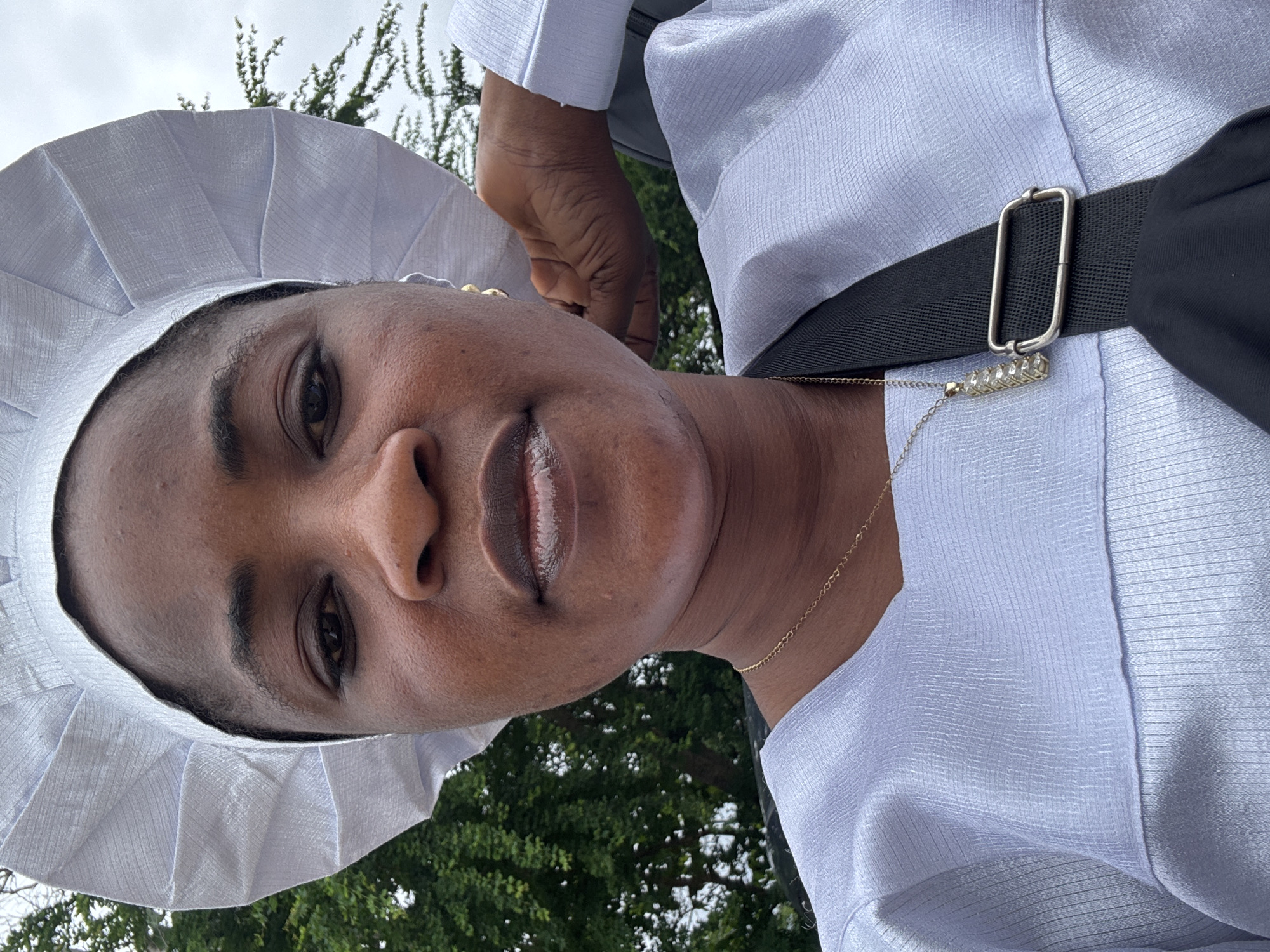 The image is a close-up, selfie-style photograph of a Black woman wearing a white, pleated head covering and a shimmering white garment. She has a gold necklace with a rectangular pendant. Her right hand is behind her head, and she is wearing a black strap across her chest. The background shows green trees and a cloudy sky, suggesting an outdoor setting. The image quality is clear, with a focus on the woman's face and upper body. There is no specific information about the location within Agaw Awusa, Nigeria.