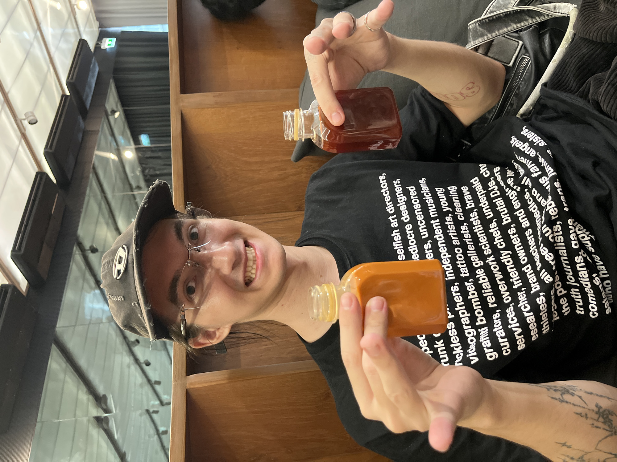 A young adult male, estimated late teens to early thirties, with long dark hair, wearing clear-rimmed glasses and a distressed dark grey baseball cap with a silver 'D' logo, is seated indoors. He wears a black t-shirt printed with white text, with legible fragments including 'selfish art directors,' 'mediocre designers,' 'videographers,' 'tattoo artists,' 'graffiti bomber,' and 'truth journalists.' He smiles broadly at the camera, displaying his teeth. A fine-line black tattoo is visible on his left forearm, and a small red tattoo is present on his right inner bicep. He holds two small, clear, square-shouldered bottles, one in each hand, both appearing uncapped. The bottle in his left hand contains a vibrant orange, opaque liquid, while the bottle in his right hand contains a dark red-brown, translucent liquid. Several rings adorn the fingers of his right hand. A black leather jacket with a zipper detail is draped to his right. The background features a light-colored wooden wall or partition behind him, a dark grey upholstered seat, and a modern interior ceiling with a reflective surface, black ventilation panels, and recessed spotlights. A green illuminated exit sign is reflected in the upper right background. Dark curtains are visible in the distant background. The scene is set within an indoor establishment, likely a cafe or lounge, in Bangkok, Thailand.