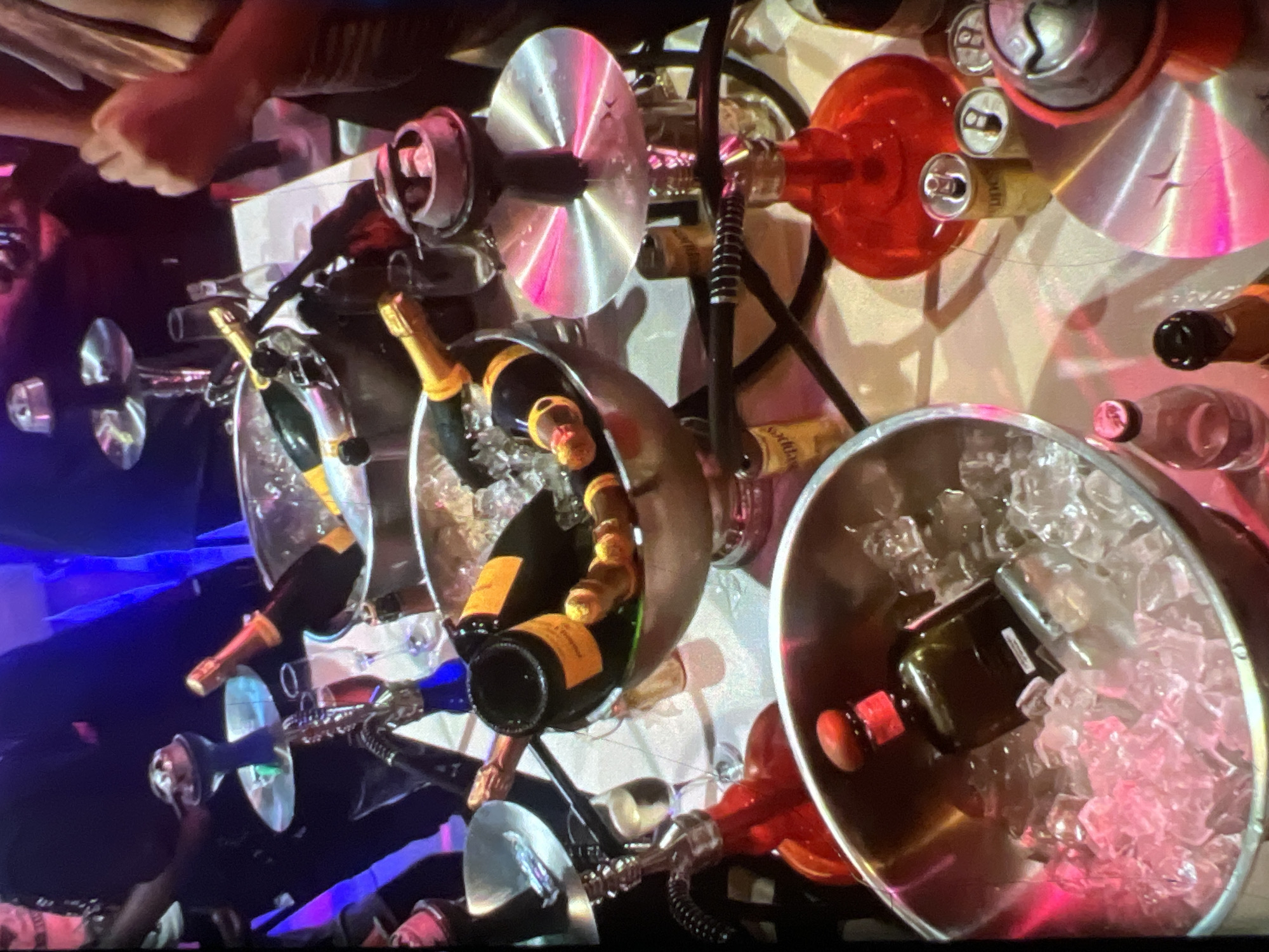 The image depicts a dimly lit indoor scene, likely a nightclub or lounge, in Lagos, Nigeria. A table is laden with various alcoholic beverages, including several bottles of champagne chilling in ice buckets. There are also shisha pipes scattered around the table, suggesting a social gathering or party. Several people are partially visible in the background, with some appearing to be standing and interacting. The ambiance is festive and celebratory, with colored lights creating a vibrant atmosphere. The focus is on the abundance of drinks and the presence of shisha, indicating a night of entertainment and indulgence.