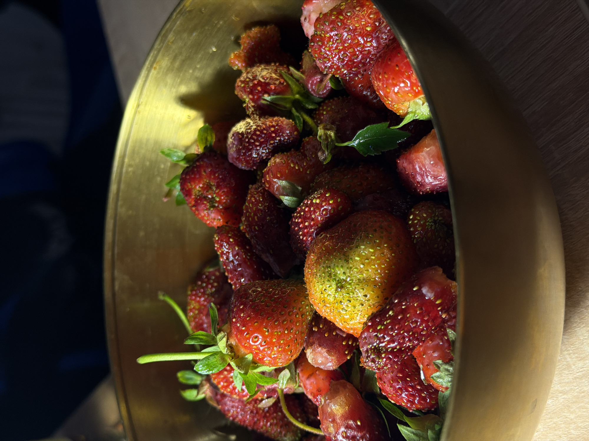 A close-up, vertical, low-angle shot captures a close-up of a brass bowl filled with fresh strawberries. The strawberries are varied in ripeness, with some exhibiting deep red hues and others displaying a mix of red and green, indicative of being partly ripe. Several strawberries have green stems and leaves attached. The bowl is resting on a wooden surface, and the background is dark and out of focus, with a hint of light suggesting natural lighting from above. The image does not depict any specific location in a city.