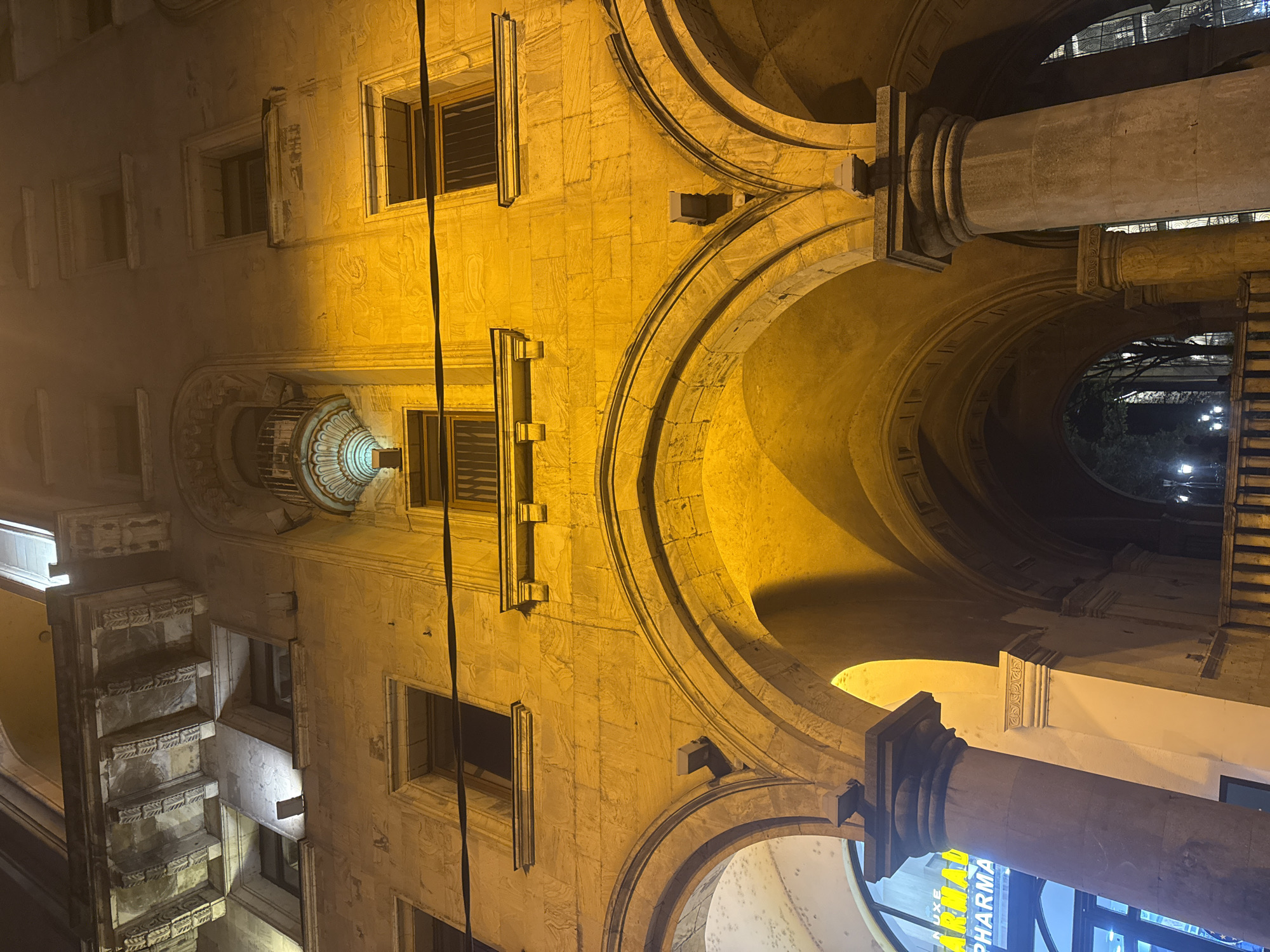 This image depicts a section of building in Tbilisi, Georgia at night. The architecture features a series of arches supported by columns, with windows and ornate stone detailing on the facade. The building is illuminated with a warm yellow light. A pharmacy storefront is visible through one of the arches.