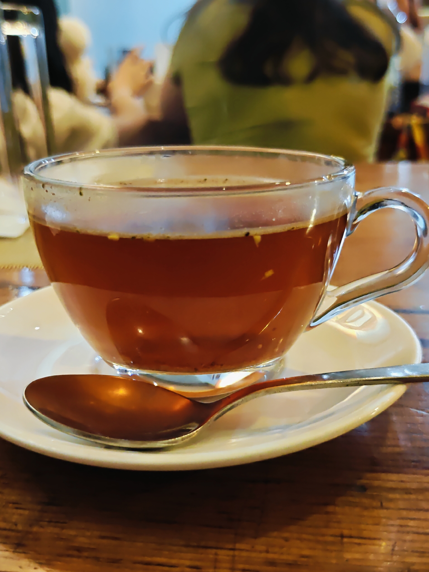 A close-up, slightly angled shot features a clear glass teacup filled with amber-colored liquid and some green specks, resting on a white saucer. A copper-colored spoon lies on the saucer in front of the cup. The teacup has a clear glass handle. The background is blurred, showing what appears to be a restaurant or cafe setting with people and muted colors, suggesting an indoor environment in Kathmandu Metropolitan City, Nepal. The table surface is made of dark wood.