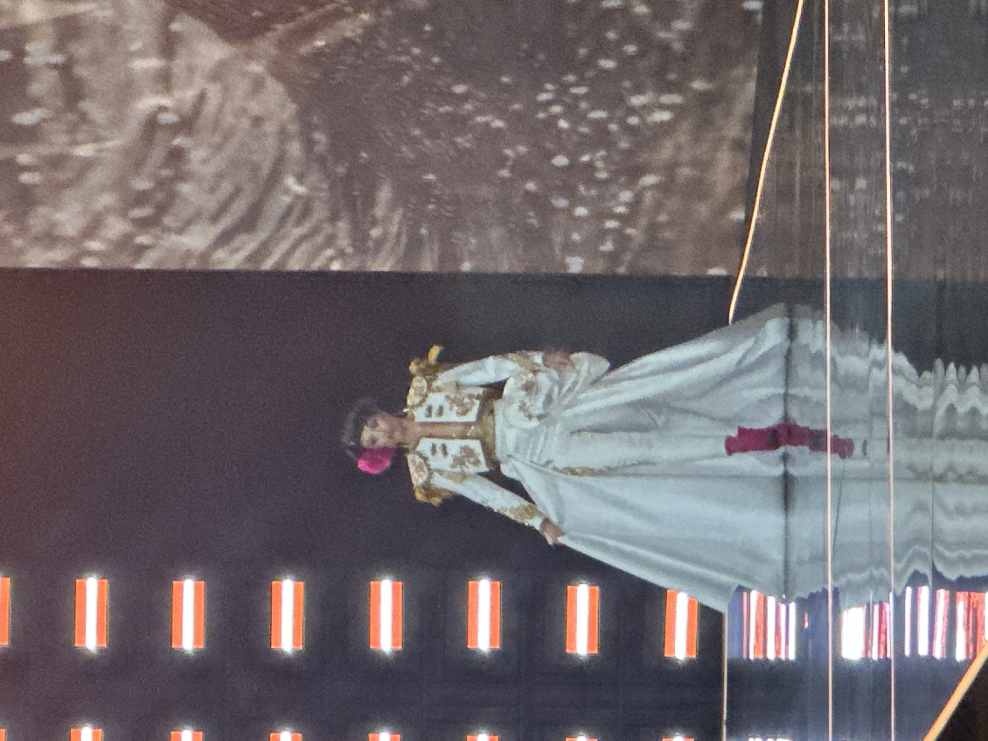 A female performer dressed as a matador stands on a stage. She is wearing a white matador costume with gold embroidery on the jacket and pants, and a large white skirt that flares out. She has a pink flower in her hair and pink boots. The stage has a dark background with rows of orange and white lights. To the right of the stage is a large textured screen displaying an abstract pattern. The floor of the stage is reflective, showing the lights and the performer's reflection. This scene is part of a performance. The location of this performance is not specified in the image.