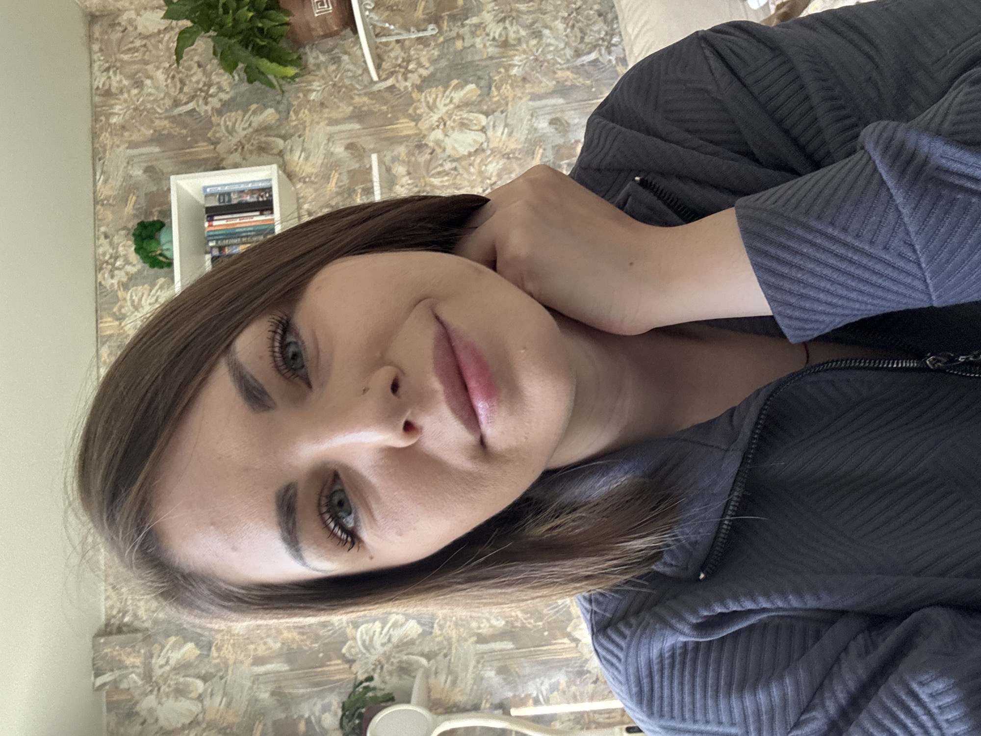 A person with shoulder-length brown hair, light skin, and blue eyes is posing indoors. They are wearing a dark-colored, textured top that appears to be a zip-up jacket or sweatshirt. Their hand is near their face, resting on the side of their head. The background includes patterned wallpaper and shelves with books and plants. The location is unknown, therefore I cannot provide a location in Раздолье, Russia.