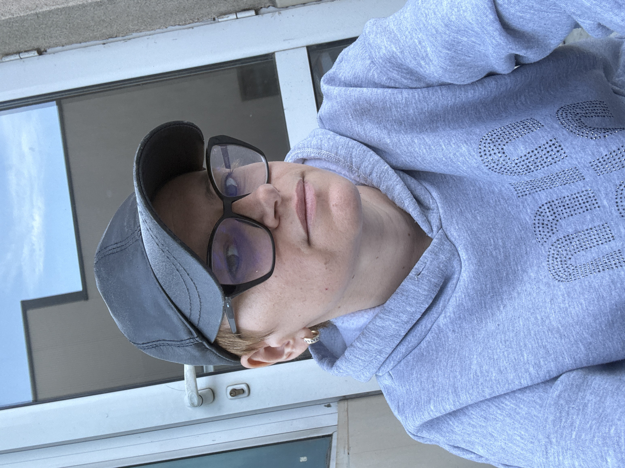 A headshot of an individual wearing a grey hooded sweatshirt with rhinestone embellishments, a dark visor, and glasses. The background shows a white window frame. The city and location are unknown.