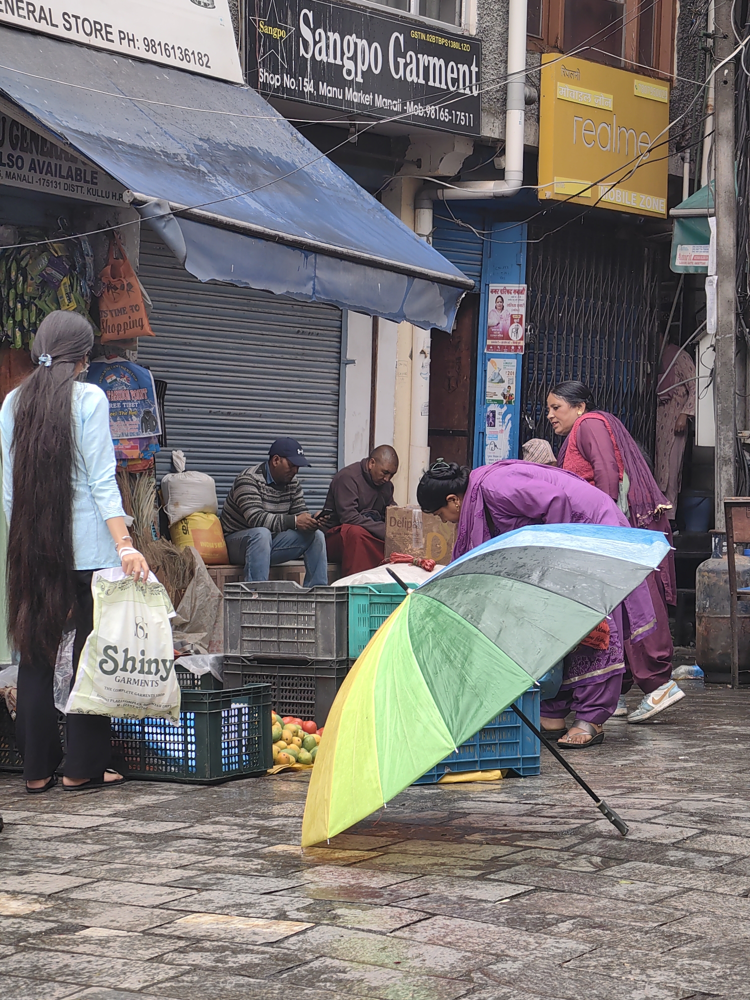The image depicts a street scene in Manali, India, likely a market area given the presence of shops and vendors. In the foreground, a large, open, colorful umbrella with sections of green, yellow, and blue is angled towards the viewer, suggesting it is being used or has recently been used for shelter. The ground is paved with stone tiles, wet from rain, reflecting the surrounding environment.
Several people are visible. On the left, a woman with very long dark hair pulled back stands facing away from the camera, holding a white shopping bag with the text "Shiny Garments" and "The Complete Garments Shop." She is wearing dark pants and sandals. In the middle ground, two men are seated. One in a striped sweater and cap is looking at his mobile phone. The other, in a maroon robe, appears to be resting.
Behind them, a stall or shopfront with a blue shutter is visible, with various items displayed, including bags and what appear to be clothing items. A sign above a shop reads "Sangpo Garment." Another yellow sign with Hindi text and the brand "realme" indicates a mobile phone shop.
Further to the right, two women are interacting. One, wearing a purple outfit, is leaning over and looking downwards. Another woman, also in a purple outfit, is standing and looking at the first woman. A large yellow gas cylinder is visible in the background. The overall atmosphere suggests a busy, perhaps rainy, market day.