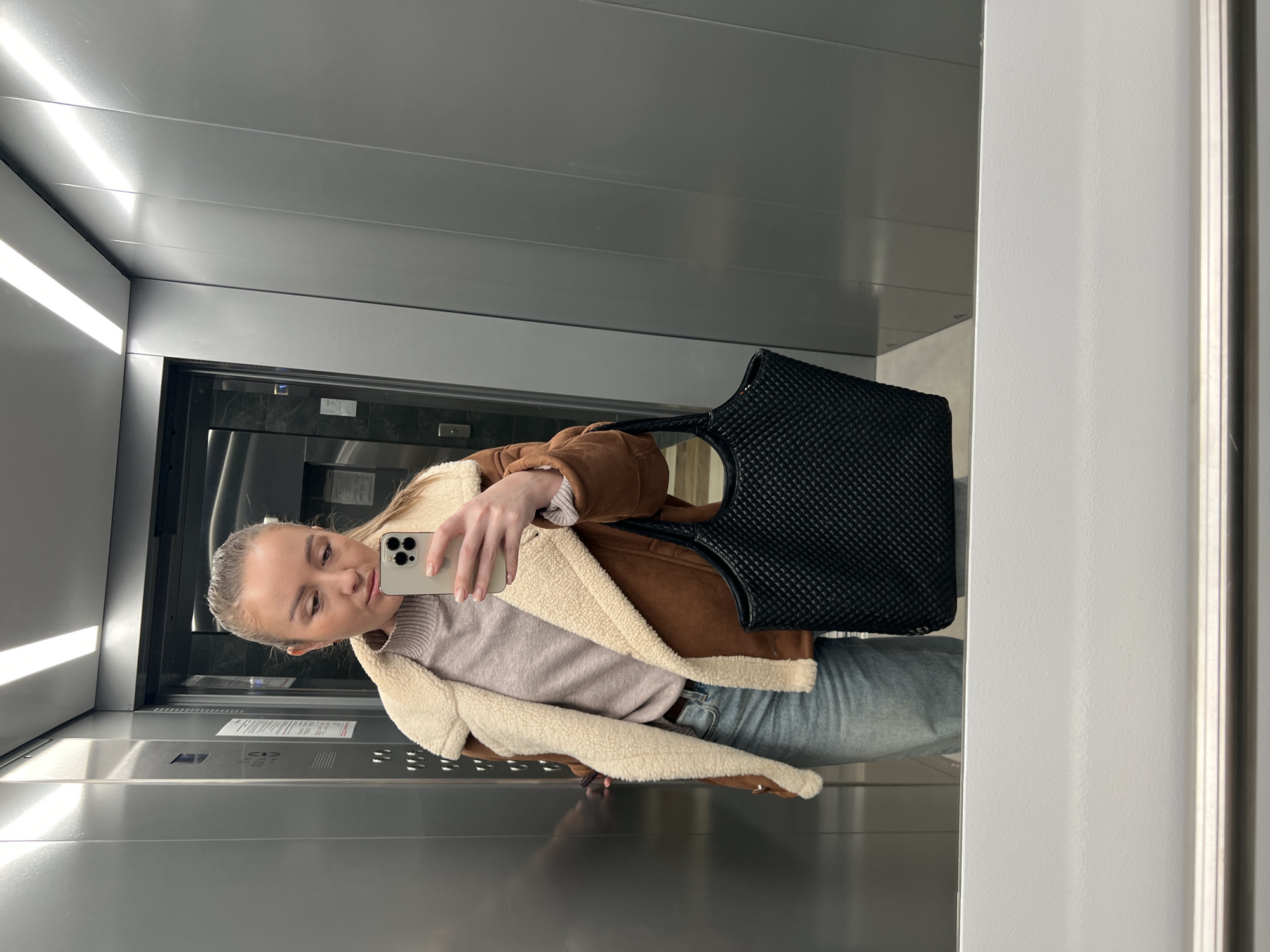 A fair-skinned adult female, appearing to be in her 20s or 30s, is depicted taking a mirror selfie inside a modern elevator. The location is an elevator car within the city of Unknown, Unknown. The subject has light brown hair pulled back, and her gaze is directed downward at a light-colored smartphone, possibly an iPhone, held in her right hand. Her fingernails are manicured with light pink polish. She is wearing a brown jacket with a cream shearling lining, a light purple ribbed turtleneck sweater, and light blue denim jeans. A dark belt is visible at her waist. A large, black, textured tote bag with a woven or quilted pattern hangs from her left arm. The elevator interior features sleek, vertically aligned gray panels on the walls and a linear light fixture overhead. A metallic elevator door with a visible control panel and placard is partially visible to the left. The bottom portion of the scene shows a reflective surface, likely the bottom of the mirror, with a white or light-colored horizontal base.
