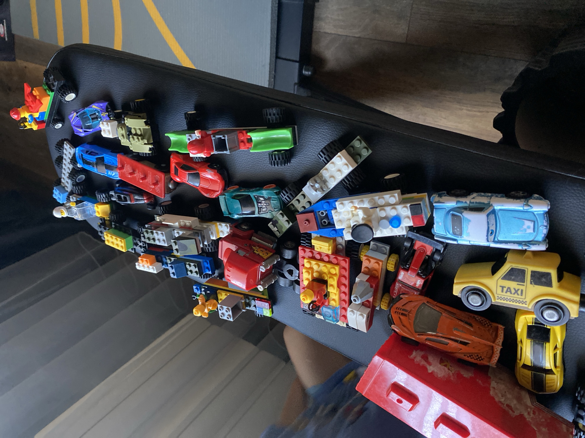 The image displays a collection of toy vehicles and building blocks, predominantly Legos, arranged on a dark, textured surface that appears to be a bench or seat. The toys are scattered and overlap, with a variety of colors and designs including cars, trucks, and figures. In the background, a grey, ribbed surface is visible, and to the right, a black object with yellow markings is present. The lighting suggests an indoor environment. The location of Зимородок Днп, Russia, is not directly indicated by any visual elements in the image.