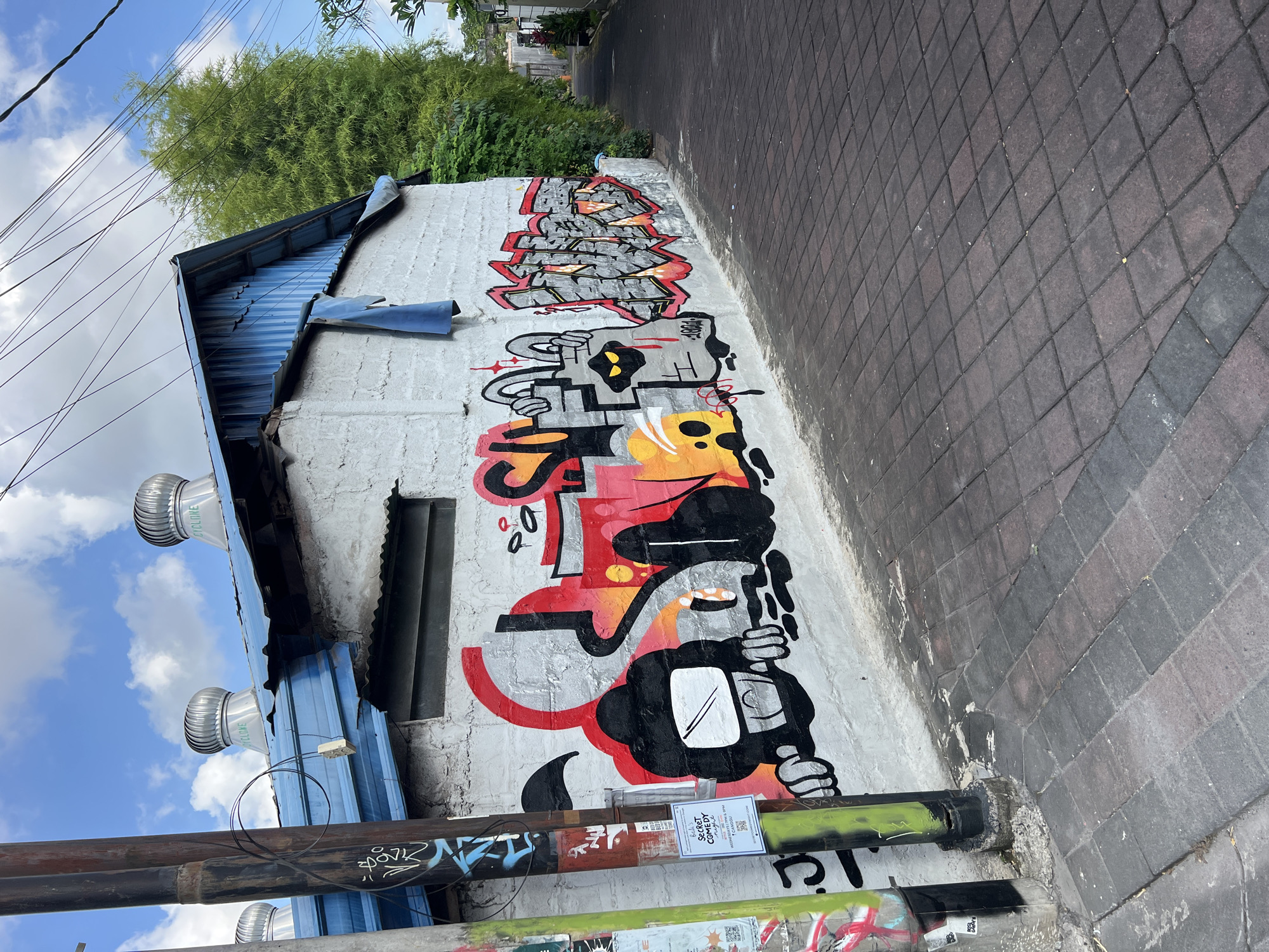 The image shows a narrow alleyway in North Kuta, Indonesia. The alley is paved with grey and black bricks. On the left is a low, white building with a blue corrugated metal roof and three silver turbine ventilators. The building's wall is covered in colorful graffiti, including stylized lettering and cartoon-like characters with black, yellow, and red accents. Below the graffiti are a series of utility pipes with additional graffiti and stickers. Above the building, trees are visible. In the background, the alley extends into the distance. The sky is visible, with a mix of blue and white clouds.