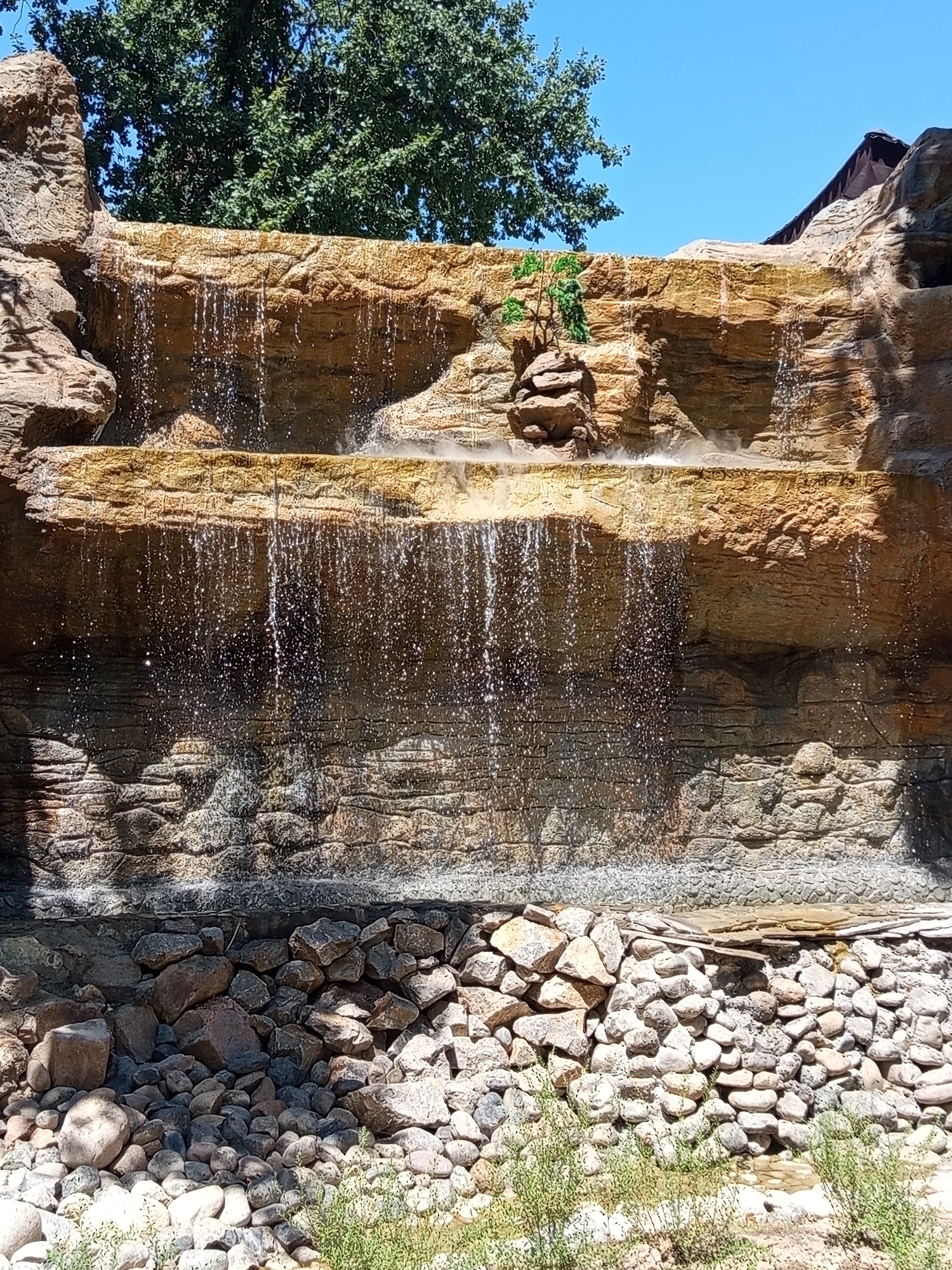 A tiered artificial waterfall is depicted, constructed from textured, rock-like material in shades of tan and orange. Water cascades down multiple levels, with a mist rising from the lower section. The top of the waterfall is partially obscured by the dark green foliage of a large tree and a portion of a dark roof structure. The base of the waterfall is a jumble of various sized stones and rocks, with some sparse green vegetation growing in the foreground. The sky above is a clear blue. The scene appears to be an outdoor installation, possibly in a park or recreational area. This waterfall is located in the zoo of the city of Shymkent, Kazakhstan.