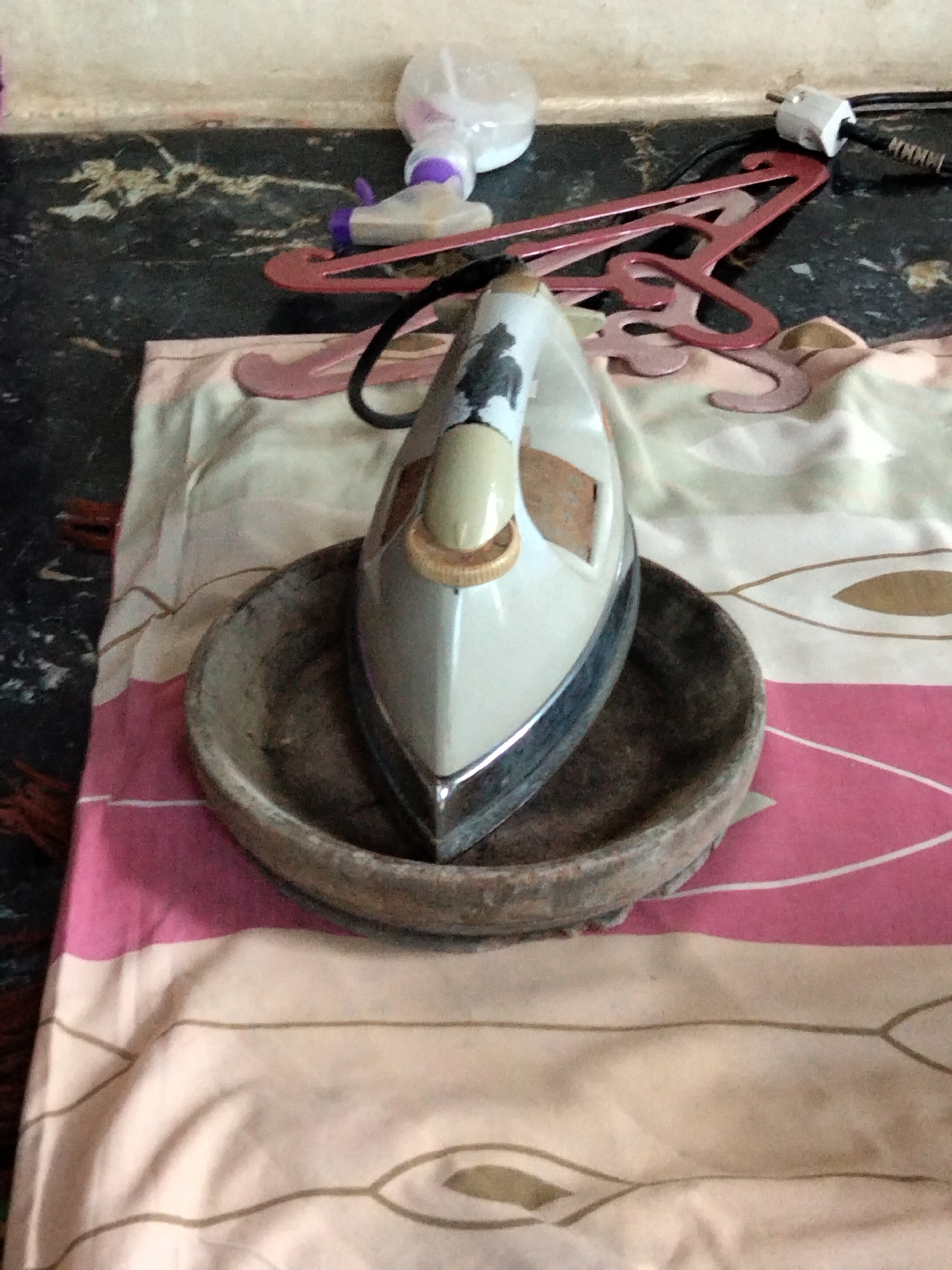 In an indoor setting, possibly a domestic dwelling in Benuaraja, Indonesia, an electric iron rests on a rustic, circular wooden dish. The iron, primarily white with a beige handle and metallic accents, shows some signs of wear, including what appears to be rust or discoloration on its upper casing. This dish is placed on a fabric, likely a tablecloth or sheet, featuring a pink and cream striped pattern with subtle, abstract designs.
In the background, a dark, speckled surface, possibly a countertop or table, is visible. On this surface are several items: a pink plastic hanger, a spray bottle with a purple nozzle and a clear plastic reservoir, and a white electrical plug attached to a black cord. The wall above the countertop is a neutral, possibly beige or off-white, and shows some minor imperfections. The overall impression is one of everyday domesticity, with the ironing equipment suggesting recent use or preparation for use.
