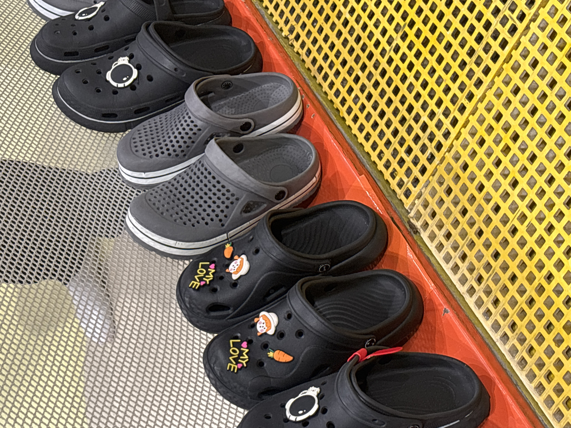A row of black and gray clogs is displayed on an orange shelf. The clogs are decorated with various charms, including carrots, cats, and a ring. The shelf is positioned above a yellow grated surface. The background consists of a mesh-like barrier. The scene appears to be in a retail environment, potentially a shoe store or a display area, possibly located in Dubai, United Arab Emirates, given the typical retail displays in the city. No people are visible in the image.