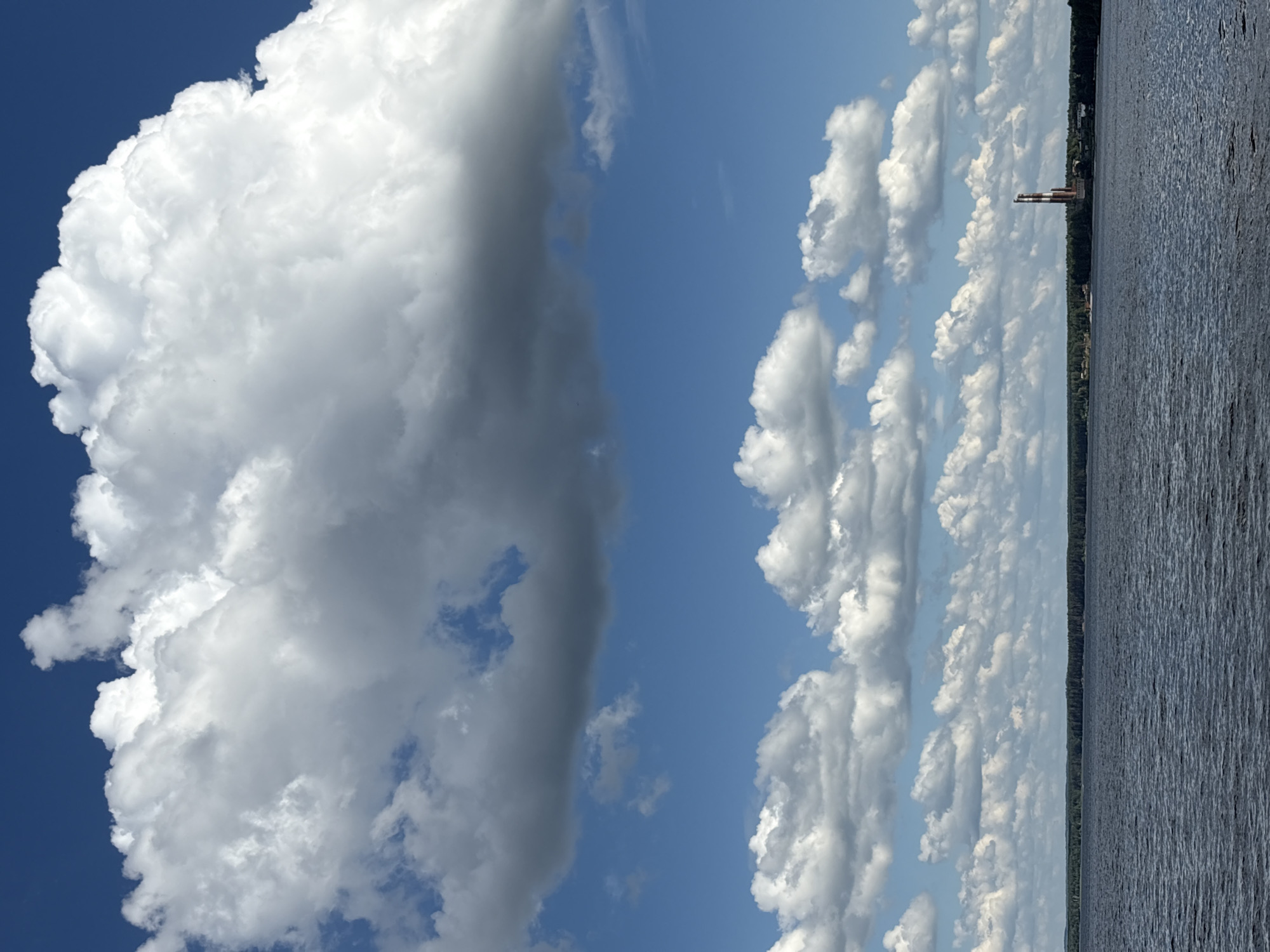 An overcast sky with large, white cumulonimbus clouds dominates the upper portion of the frame, with patches of bright blue sky visible between them. A second layer of smaller, white cumulus clouds stretches across the horizon. Below the clouds, a wide, dark blue body of water, possibly a river or reservoir, fills the lower half of the image. The water's surface is rippled, indicating a gentle breeze. In the distance, a dark green line of trees forms the shoreline, interrupted by a red and white smokestack of a factory. The smokestack is tall and slender, with white bands alternating with red sections. The overall scene depicts a tranquil, natural landscape with a prominent industrial element on the horizon, likely in the city of Chaikovsky, Russia, known for its Kama River and surrounding industrial facilities. There are no people or animals visible in the image.