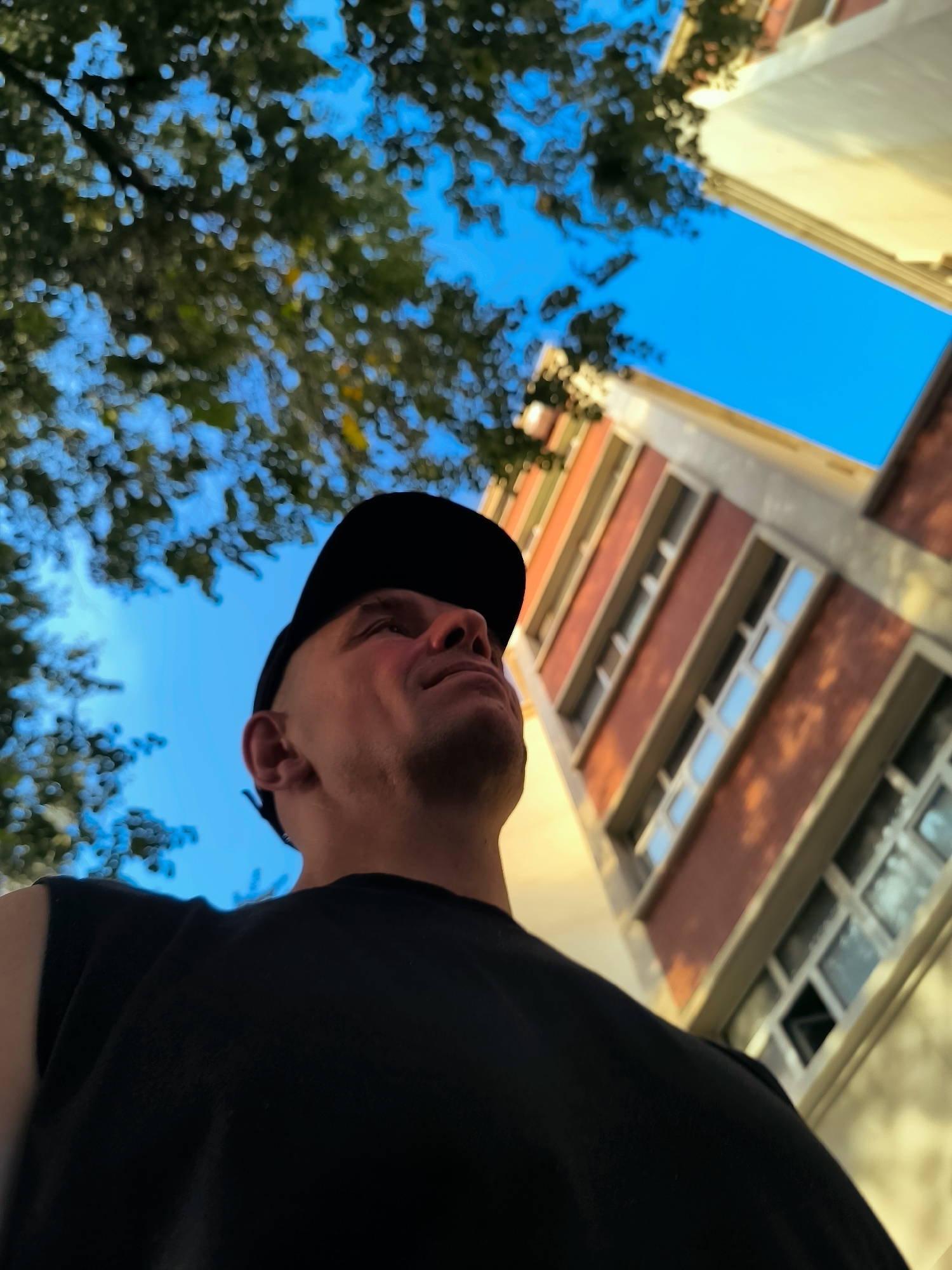 An upward-angle, medium shot captures a person wearing a black baseball cap and a black t-shirt against a bright blue sky and a building. The person's head is tilted upwards, their gaze directed towards the sky. The person's right ear is visible. A portion of a multi-story building with red brick and beige trim is seen on the right side of the frame. Lush green foliage is visible in the upper left corner. The scene is set in the city of Ürümqi, China.
