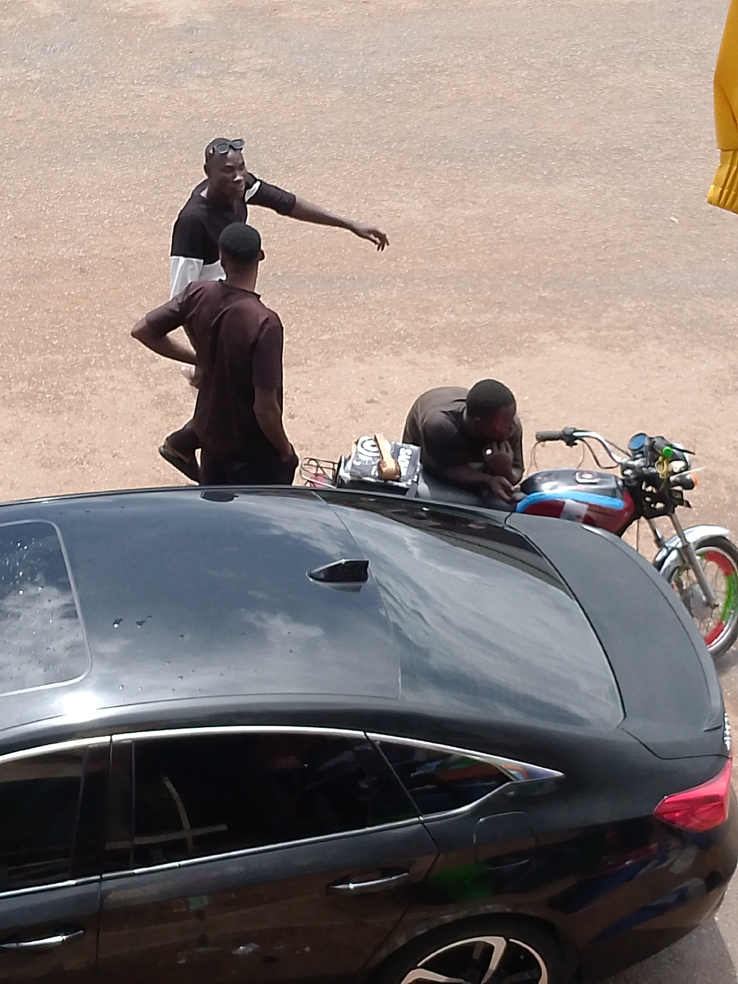 In Bukuru, Nigeria, a daytime scene unfolds on a gravelly, light-colored road. Two men stand near a black sedan. The man on the left wears a black shirt with a white stripe across the chest and dark sunglasses perched on his head. He is gesturing with his right arm, pointing away from himself, and appears to be interacting with another man partially obscured behind him, who is wearing a dark brown short-sleeved shirt and dark pants. A motorcycle with a multi-colored stripe on its fuel tank is parked behind the sedan. A third man, wearing a dark short-sleeved shirt, is leaning on the motorcycle, resting his chin on his hands. The black sedan occupies the foreground of the image, angled to the right, with its rear end closer to the viewer. The road surface is visible around the vehicles and people, with a lighter-colored texture suggesting gravel or compacted earth.