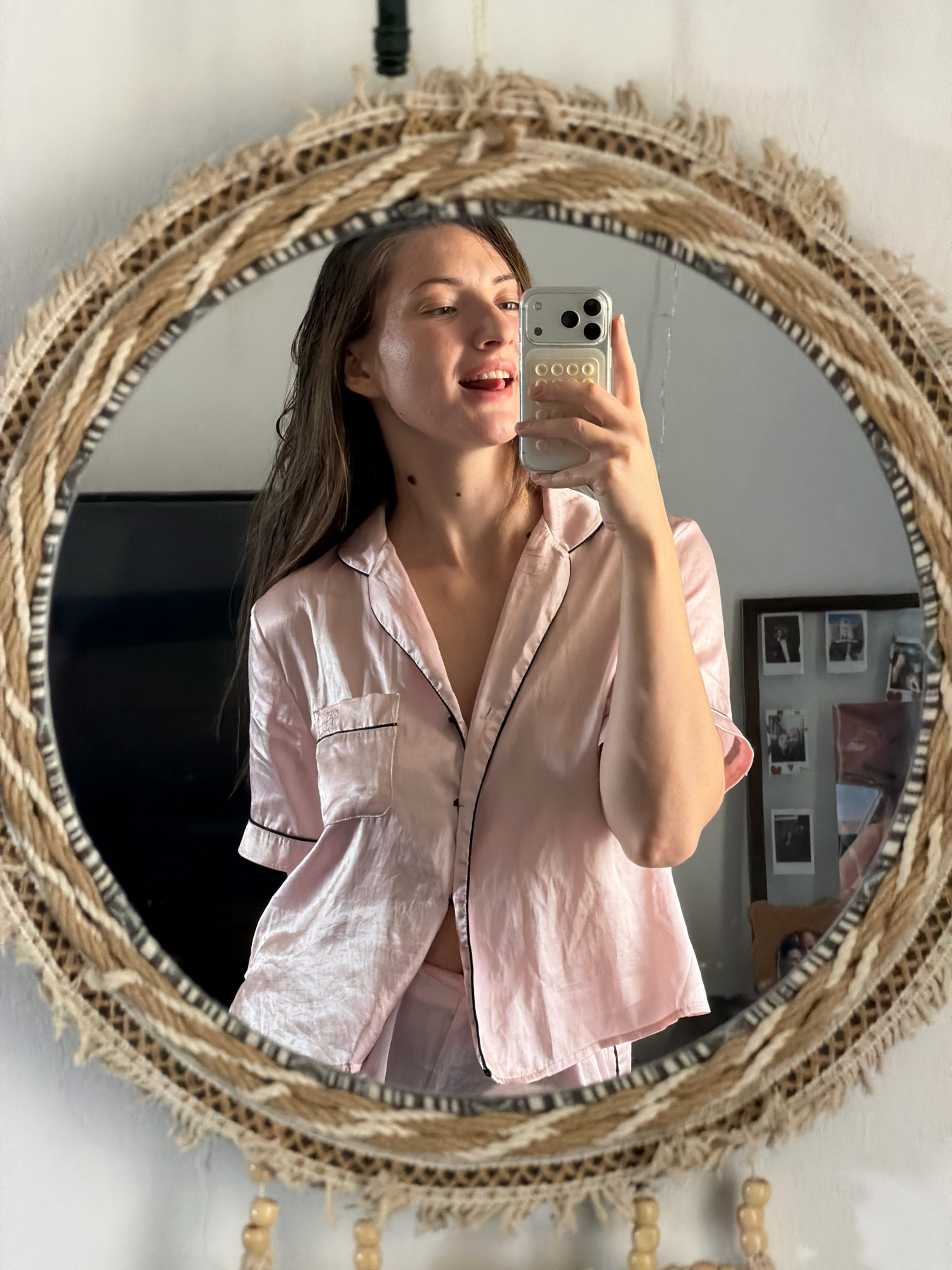 A young woman with long, dark hair takes a mirror selfie in what appears to be a bedroom. She is wearing a light pink satin pajama set with black piping. She is holding a silver smartphone in her right hand and is sticking her tongue out playfully. The mirror has a unique, circular frame made of woven materials, possibly straw or rope, with a fringed edge. In the background, reflected in the mirror, a black television is visible, and a framed collage of photographs hangs on the wall. The overall impression is casual and domestic. The location is identified as Sharm El Sheikh, Egypt.