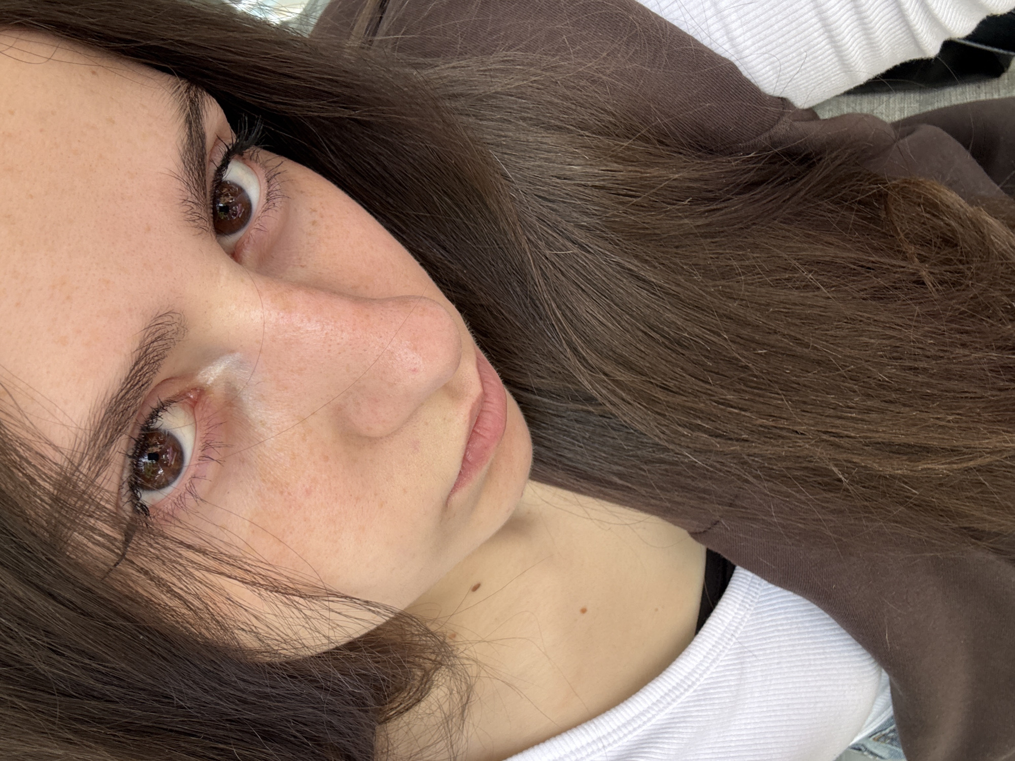 A close-up, tilted selfie of a fair-skinned young woman with freckles and brown eyes. Her brown hair is long and partially obscures her face. She is wearing a white top and a brown hooded sweatshirt. There is no indication of location in the city of Yerevan, Armenia.