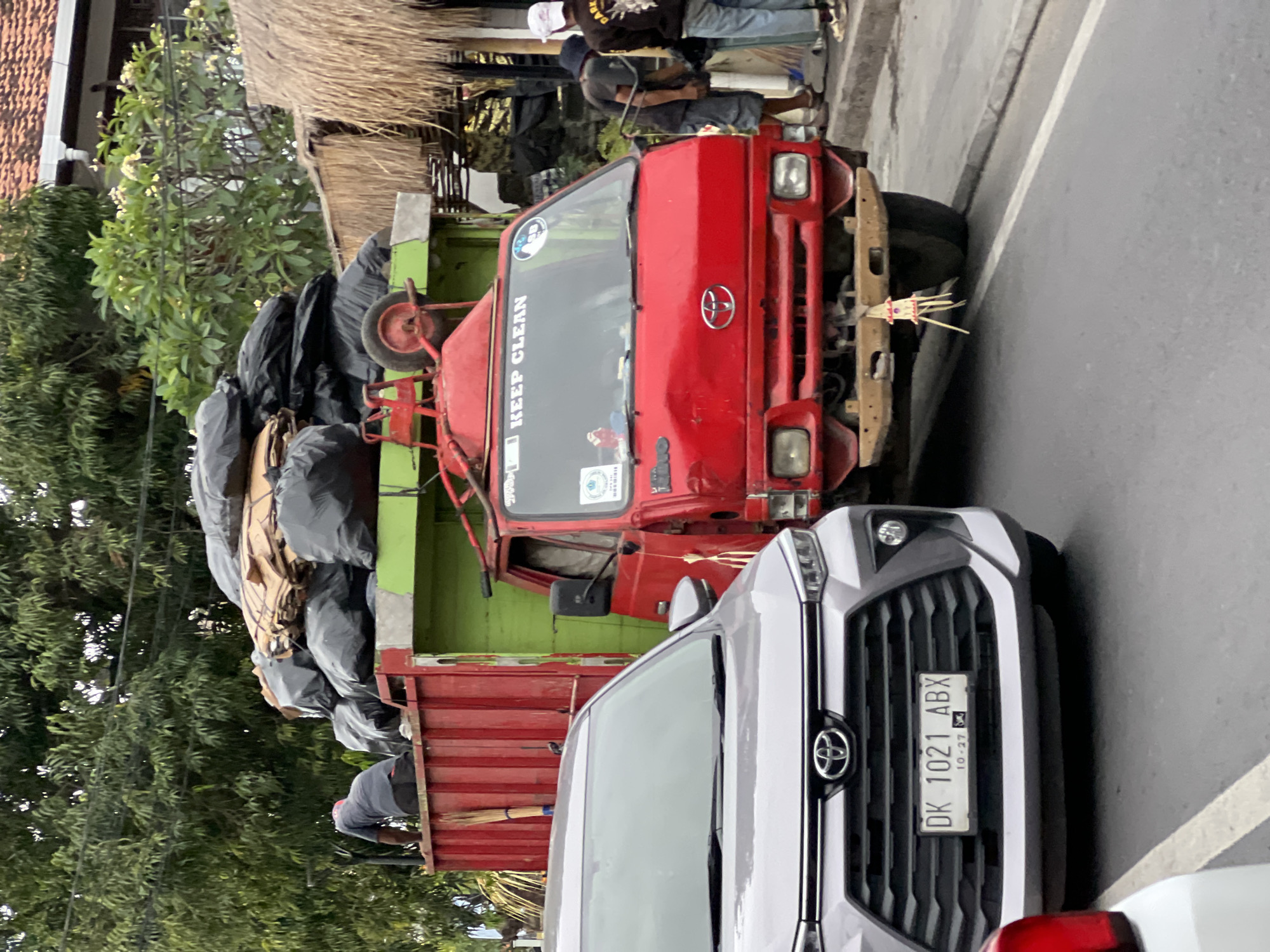 A red and green garbage truck is parked on a street in Denpasar, Indonesia. The truck is heavily laden with black garbage bags and cardboard boxes. A person is visible in the back of the truck, appearing to be arranging the load. In front of the garbage truck, a silver Toyota car is parked, partially obscuring the garbage truck's license plate. The car's license plate reads "DK 1021 ABX". Two men are standing near the front of the garbage truck, on the sidewalk. The environment appears to be a street in a tropical location, with trees and buildings visible in the background. The sky is overcast.