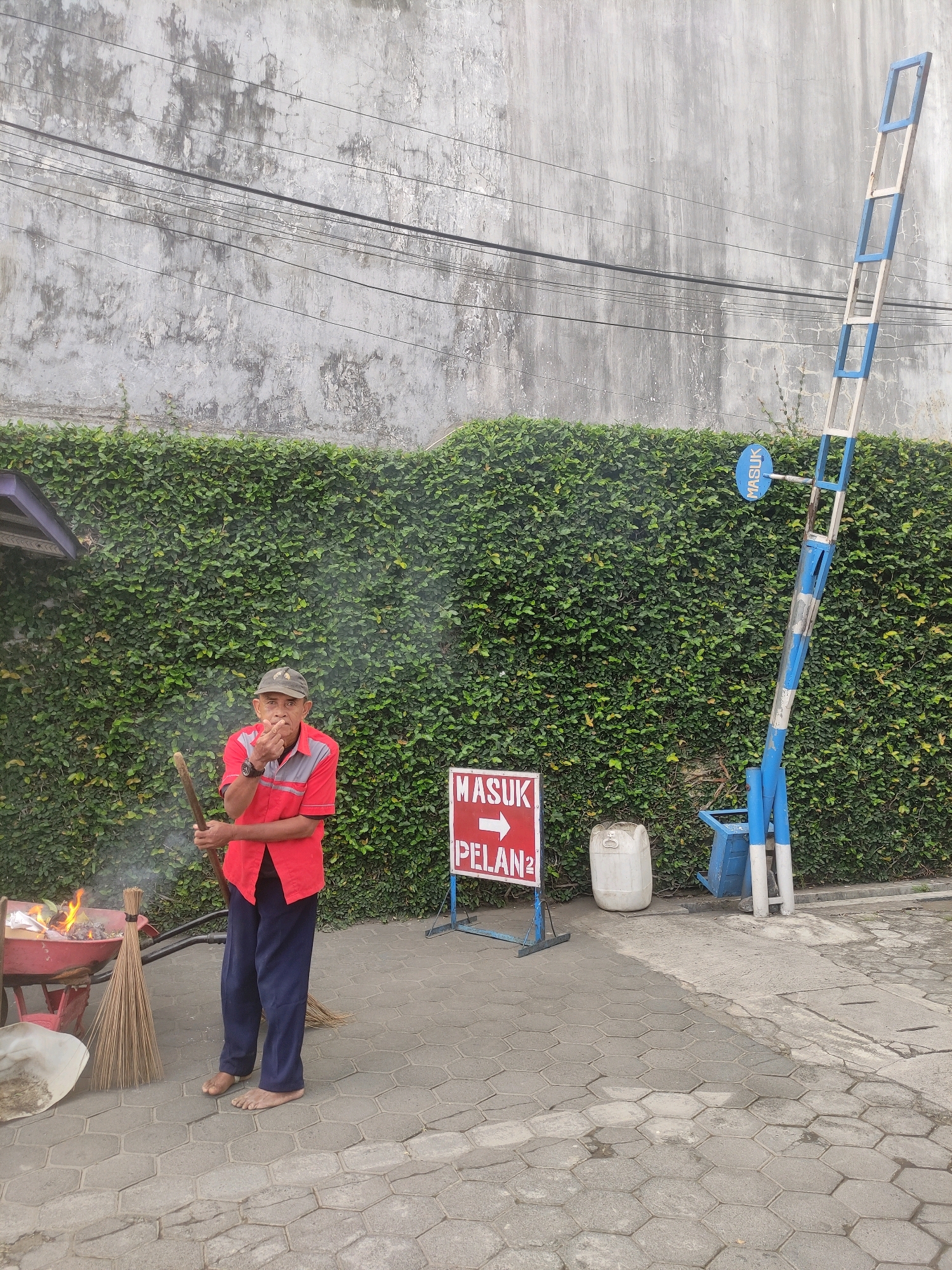 In Genteng, Indonesia, an elderly man in a red shirt and dark pants stands barefoot on a paved surface, holding a broom and a stick. He is near a burning pile of leaves in a red wheelbarrow, with smoke rising into the air. To his right, a white plastic jug and a blue and white striped barrier gate stand against a wall covered in green ivy. A red and white sign on a stand reads "MASUK PELAN" with an arrow pointing to the right, indicating "Enter Slowly". Overhead, electrical wires are visible against a grey, weathered wall.